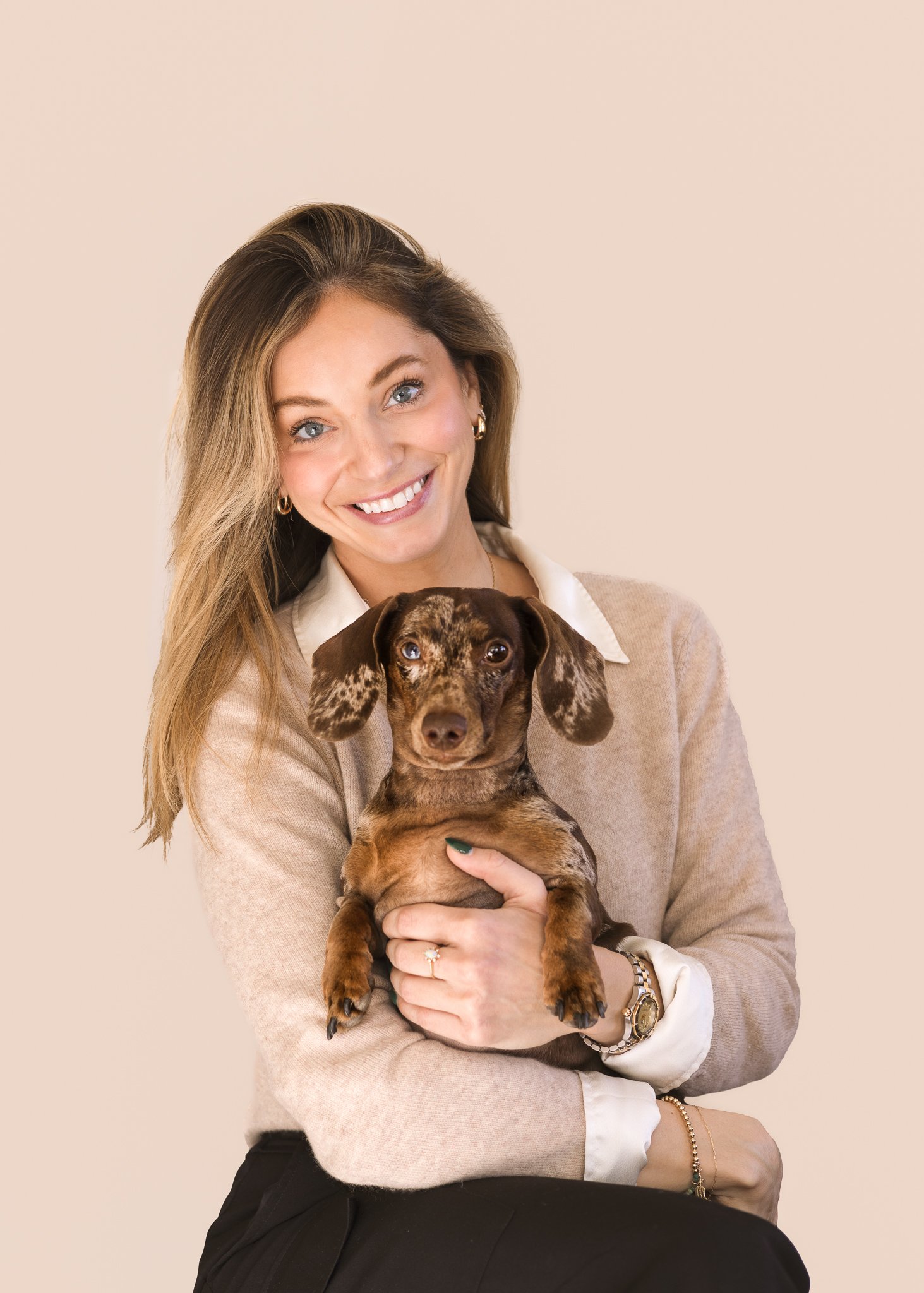 A woman with long blonde hair smiling while holding a brown and tan dapple dachshund puppy against a beige background.