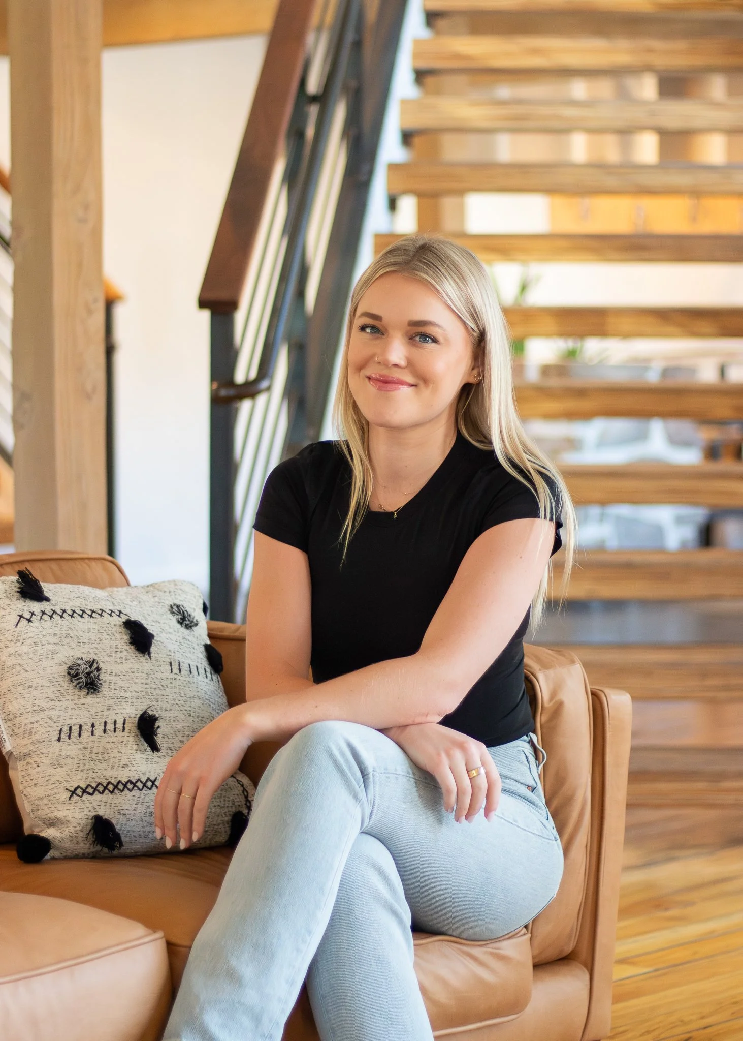 A young woman with blonde hair sitting on a tan leather sofa, smiling at the camera, in a cozy, modern home interior with wooden stairs and a pillow nearby.