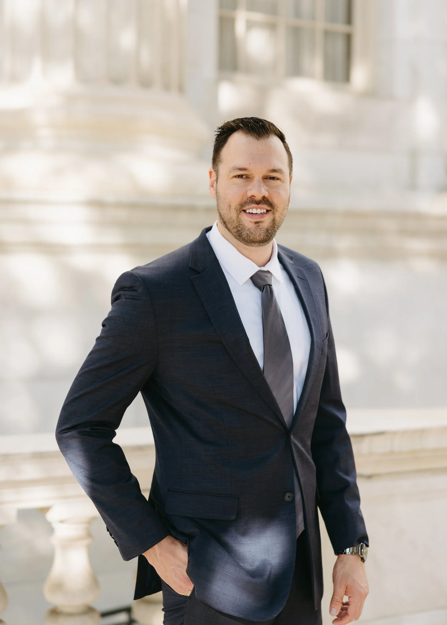 A man in a dark suit and tie standing in front of a marble staircase with balustrades.