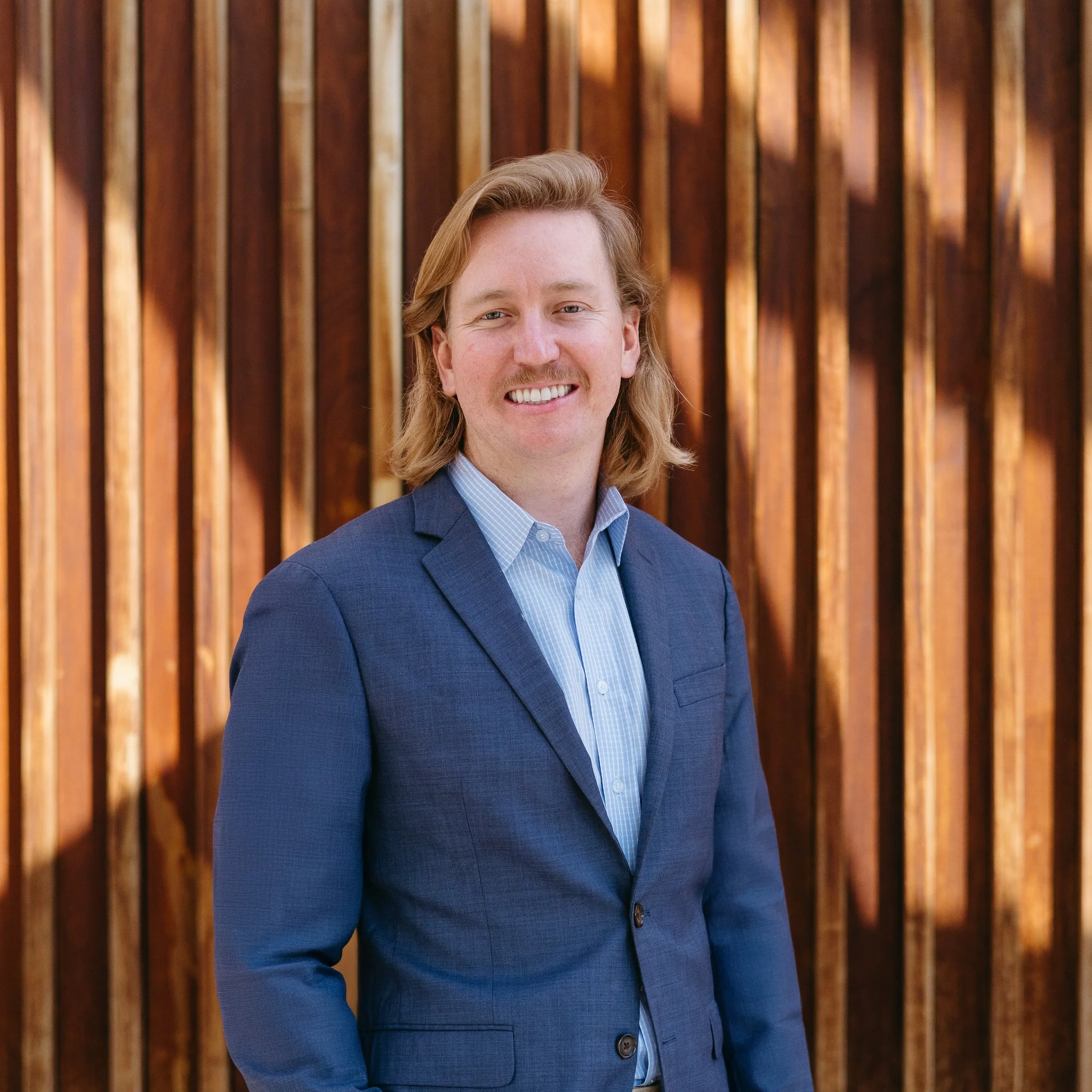 A man with shoulder-length blonde hair, a mustache, wearing a blue suit jacket and light blue shirt, standing in front of a wooden background.