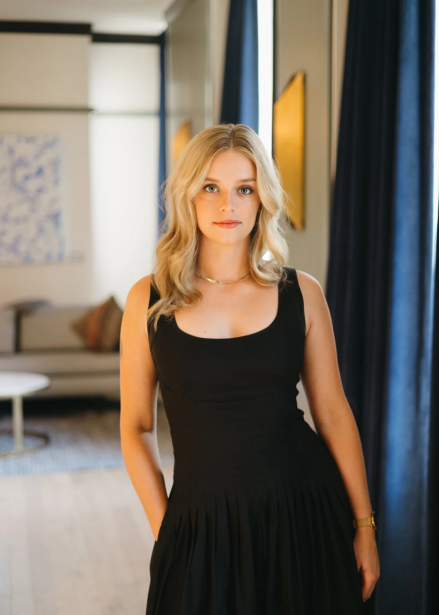 A young woman with blonde hair in a black sleeveless dress standing in a well-lit room with blue curtains, artwork on the wall, and a sofa in the background.