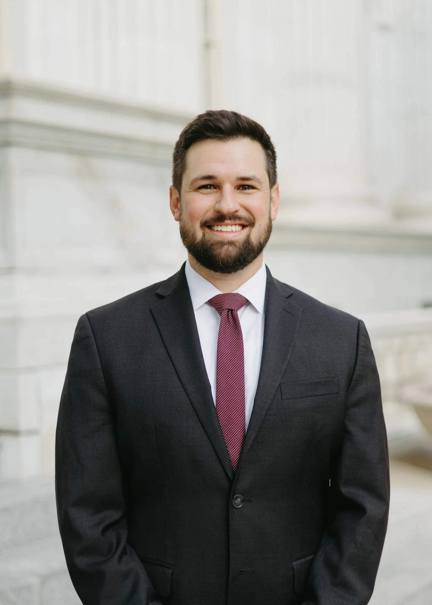 A smiling man in a black suit, white shirt, and red tie standing in front of a white marble building with stairs.