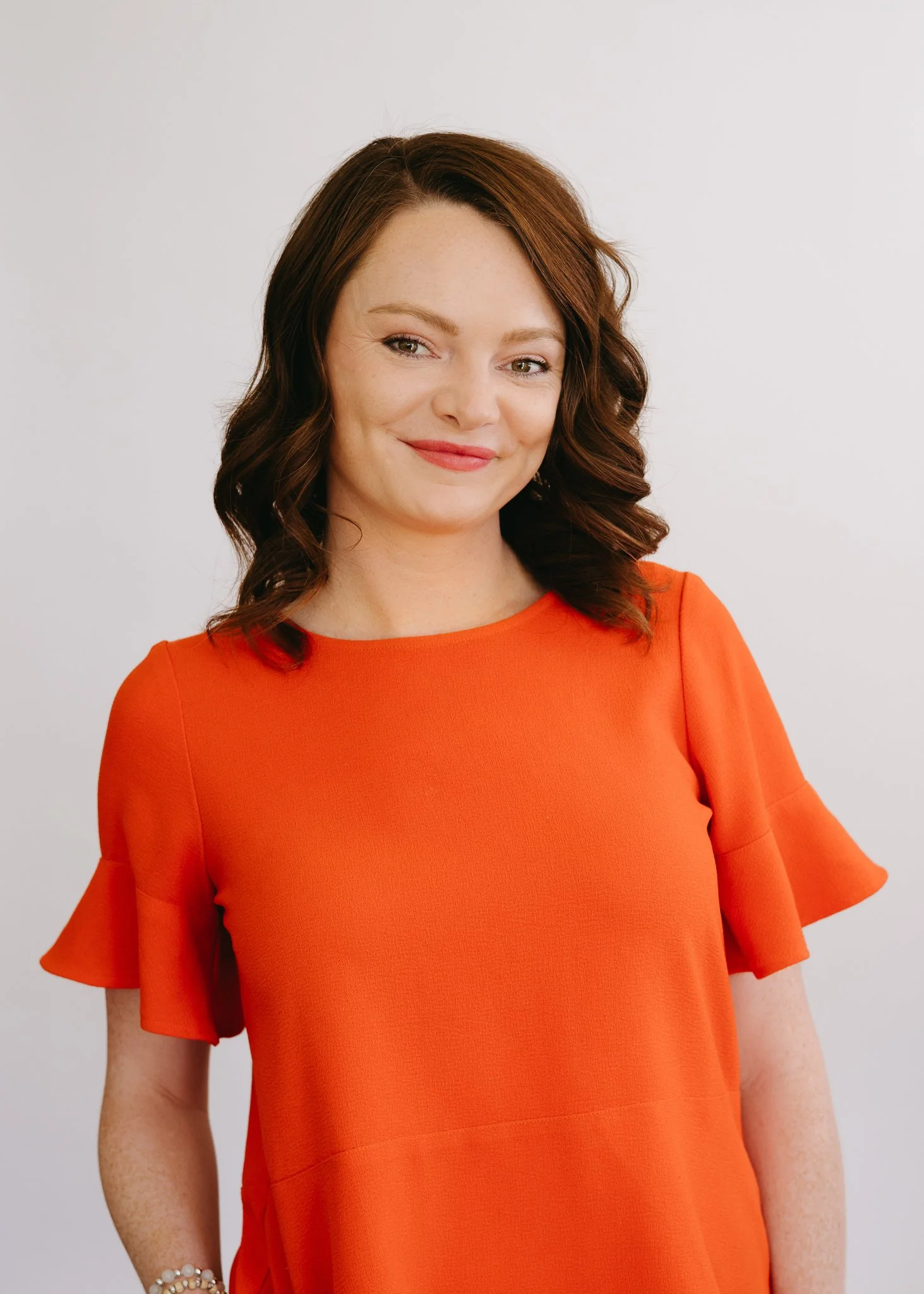 A woman with shoulder-length dark red hair, wearing a bright orange dress with ruffled sleeves, smiling and standing against a plain white background.