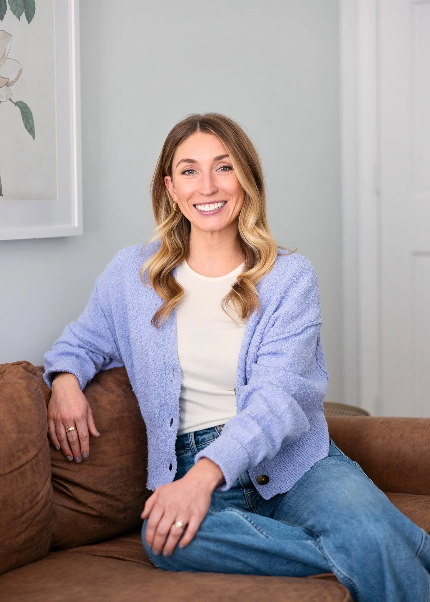 A woman with blonde wavy hair, wearing a white shirt, blue cardigan, and jeans, sitting on a brown couch and smiling at the camera.