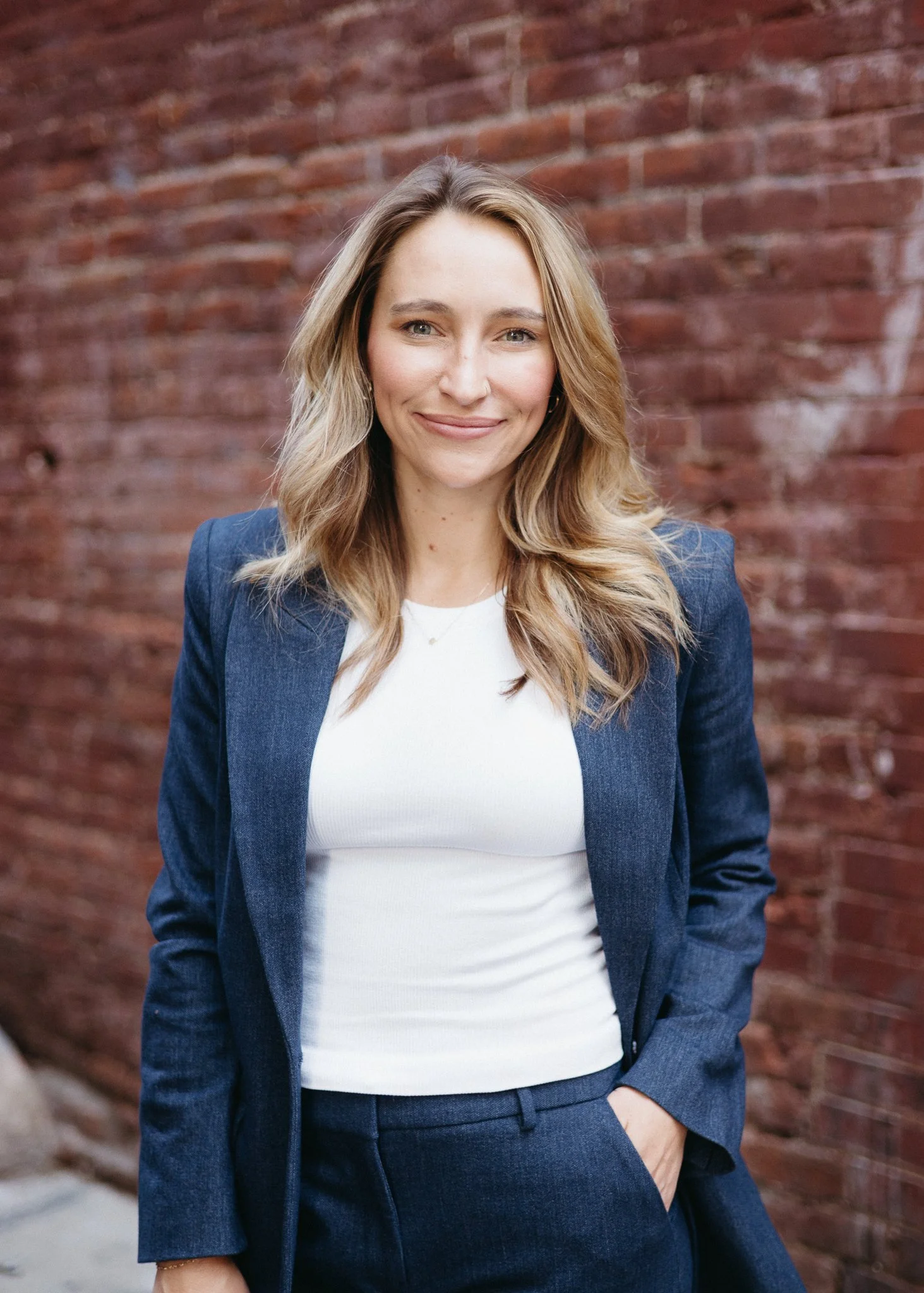 A woman with long wavy blonde hair, wearing a navy blazer and white top, standing against a red brick wall, smiling with one hand in her pocket.