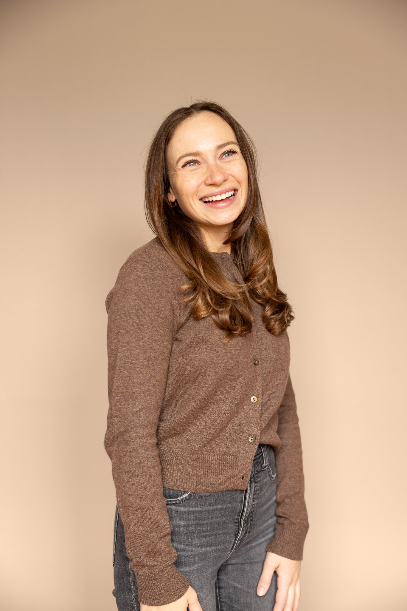 A young woman with long brown hair, smiling with teeth showing, standing against a beige background, wearing a brown sweater and gray jeans.