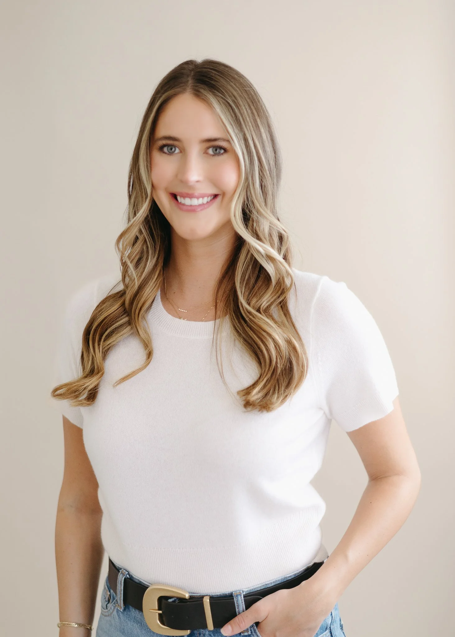 A young woman with long wavy blonde hair, blue eyes, wearing a white short-sleeve shirt, a black belt with a gold buckle, and blue jeans, smiling against a plain, light-colored background.