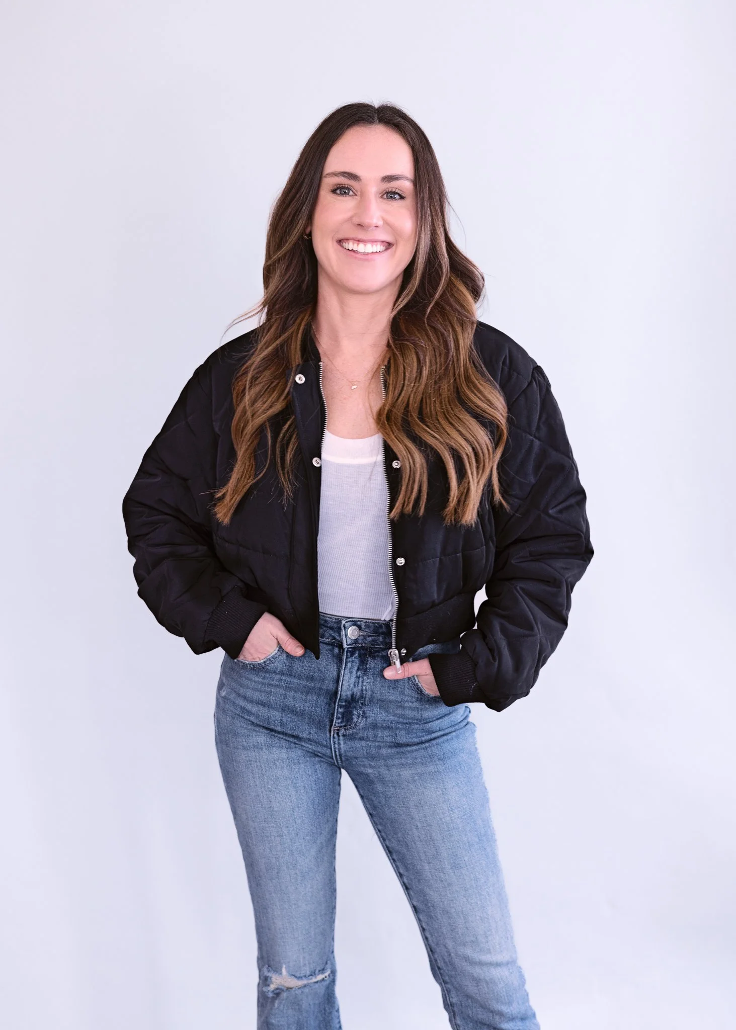 A woman with long wavy brown hair smiling, wearing a black jacket over a white top and ripped blue jeans, standing against a plain light background.