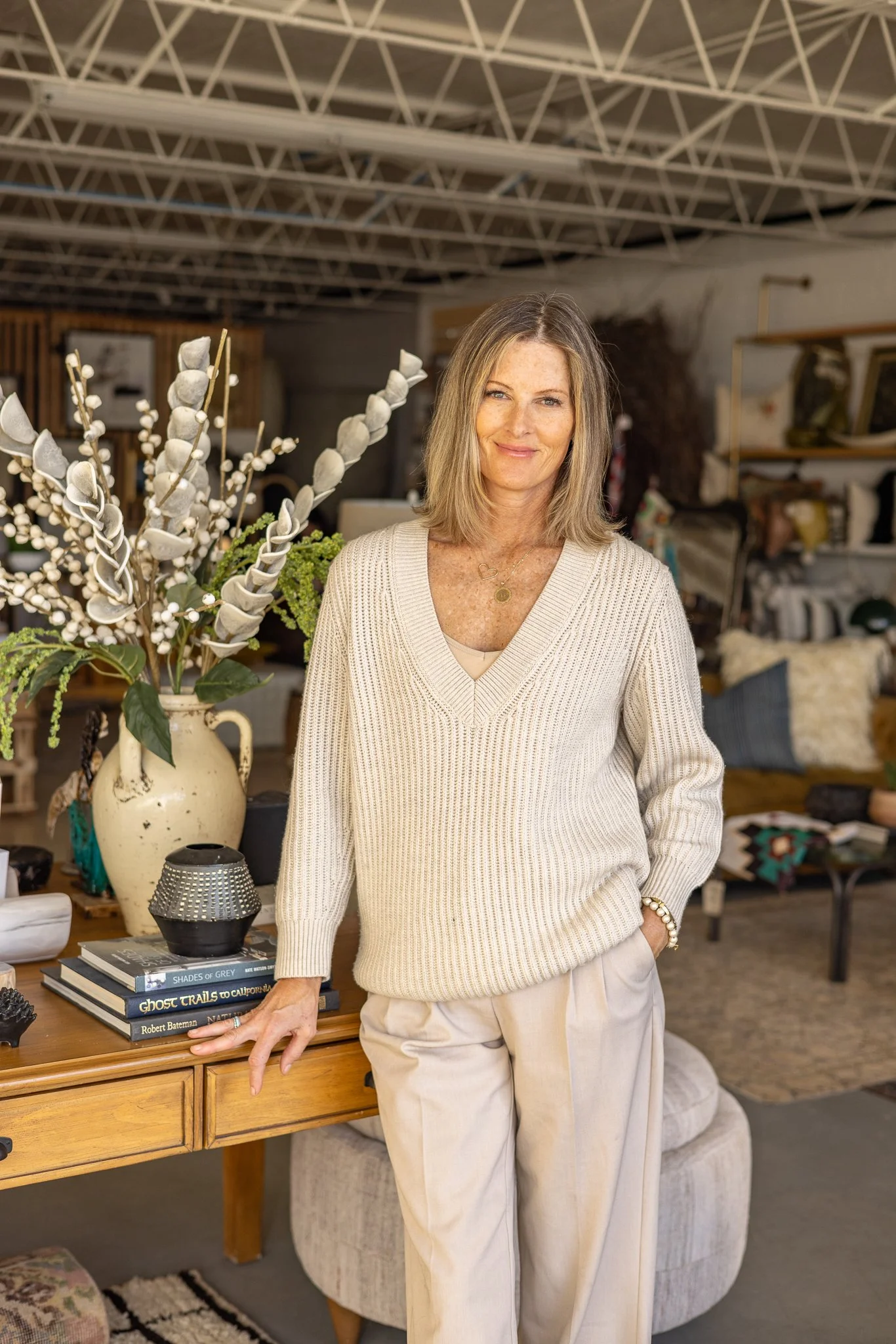A woman with blonde hair standing indoors in a cozy, styled room with decorative books, a large vase with white artificial flowers, and comfortable furniture in the background. She is wearing a cream-colored sweater and light-colored pants, smiling s