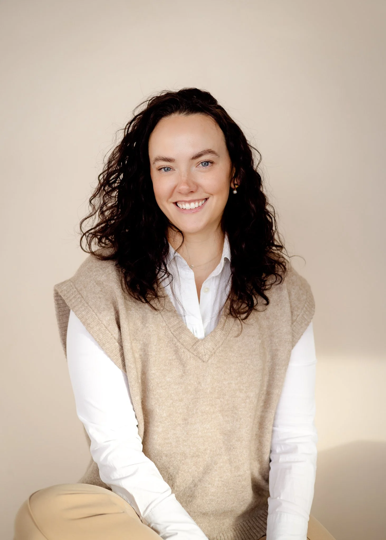 Smiling woman with curly dark hair, wearing a beige sweater vest over a white button-up shirt, sitting against a light, neutral background.