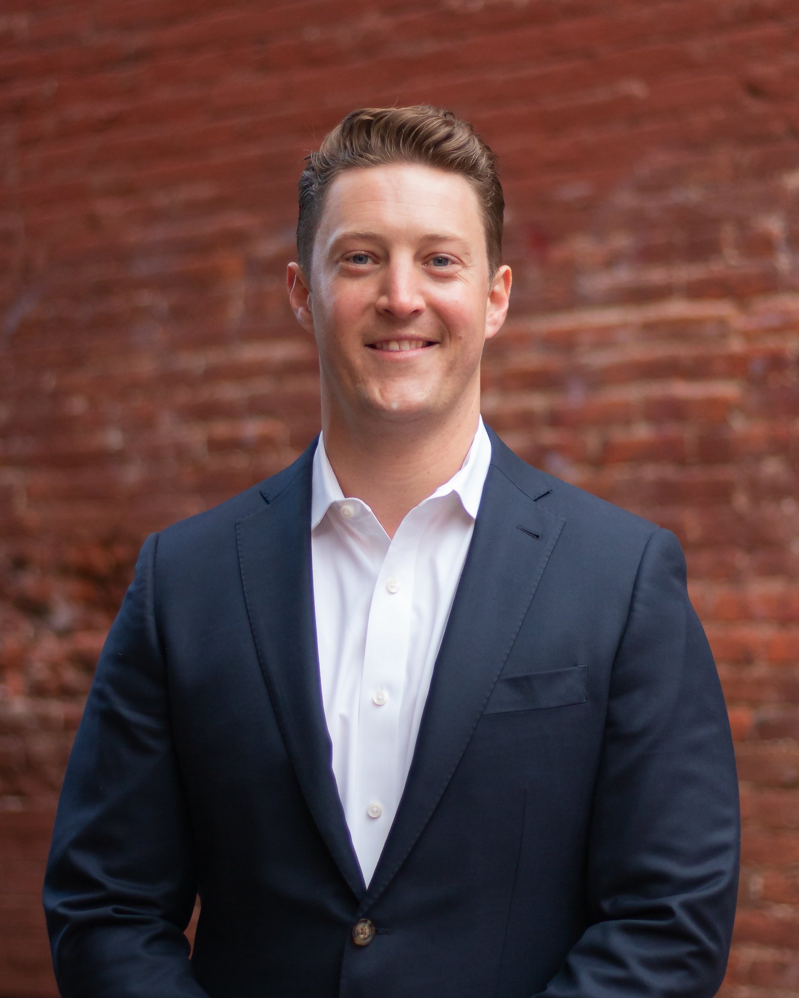 A young man in a navy blue suit and white dress shirt standing in front of a red brick wall, smiling at the camera.