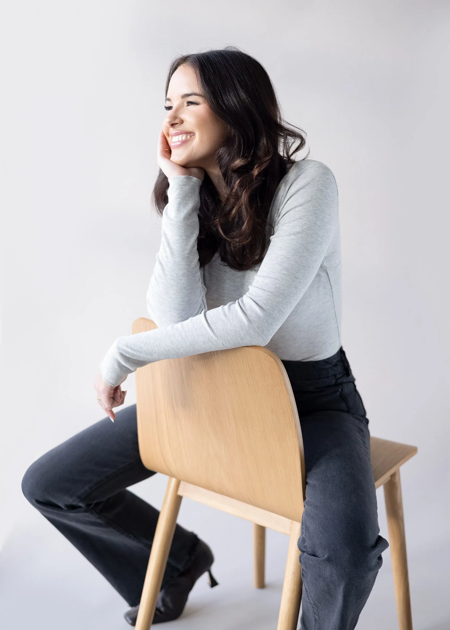 A woman sitting on a wooden chair, smiling with her hand resting on her face, wearing a gray long-sleeve shirt and black jeans.