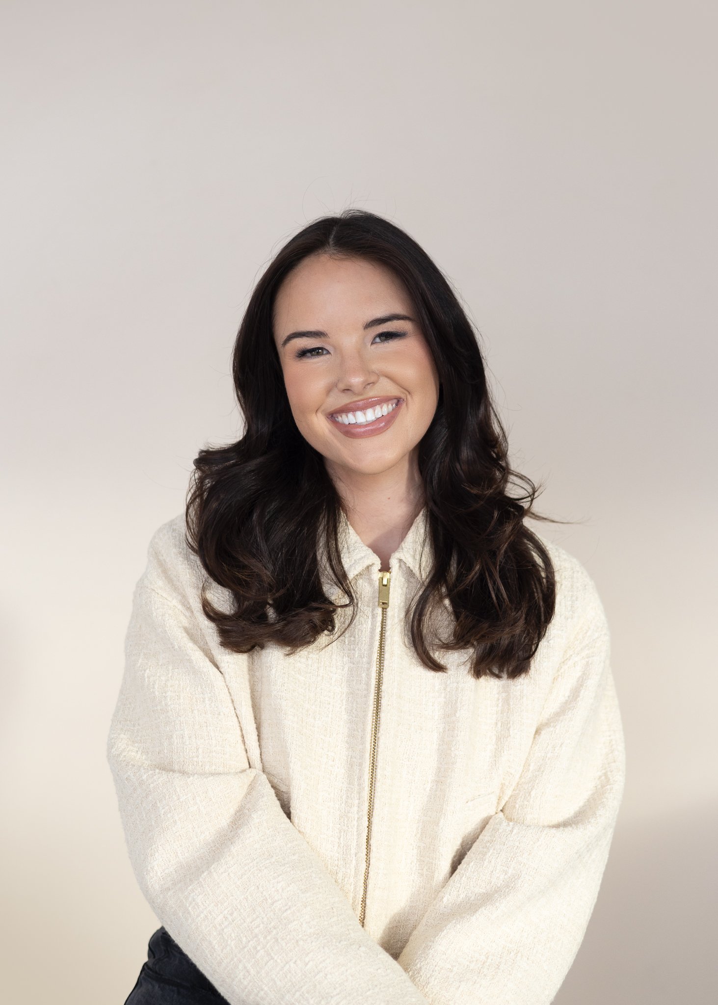 Smiling young woman with dark wavy hair, wearing a cream zip-up jacket, against a plain light background.