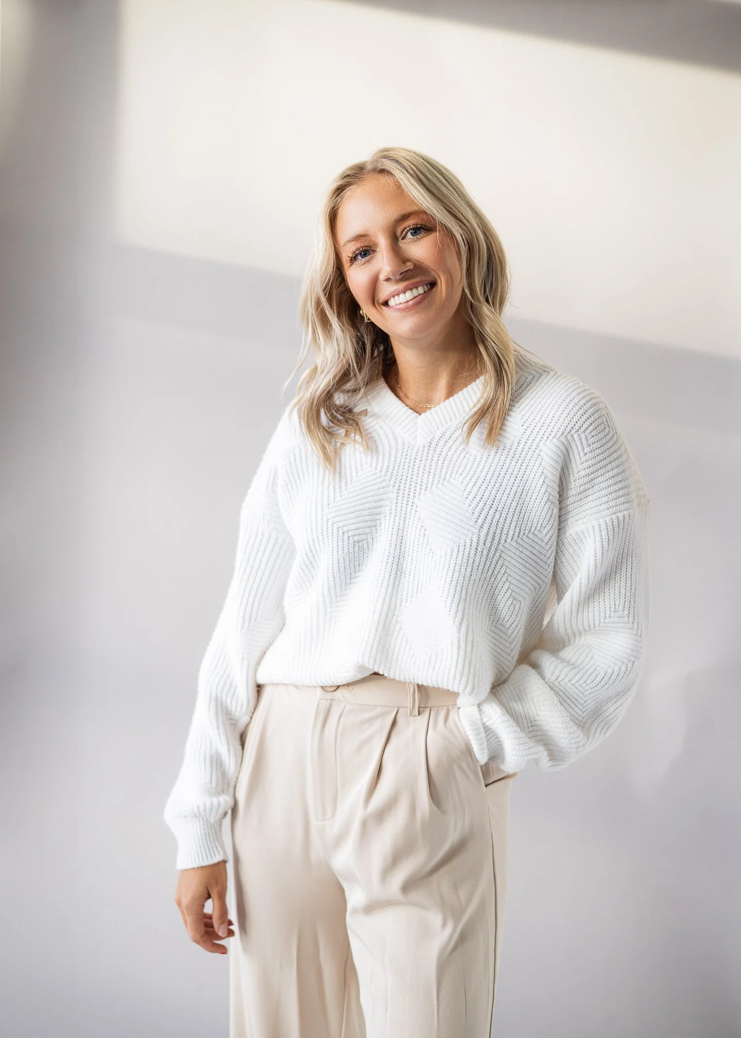 A woman with blonde wavy hair smiling, wearing a white sweater and beige pants, standing indoors against a plain light background.
