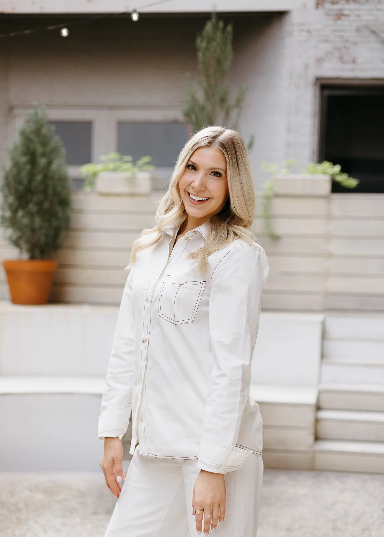 A smiling woman with blonde hair wearing white pajamas standing outdoors in front of a backyard with potted plants and a wooden fence.