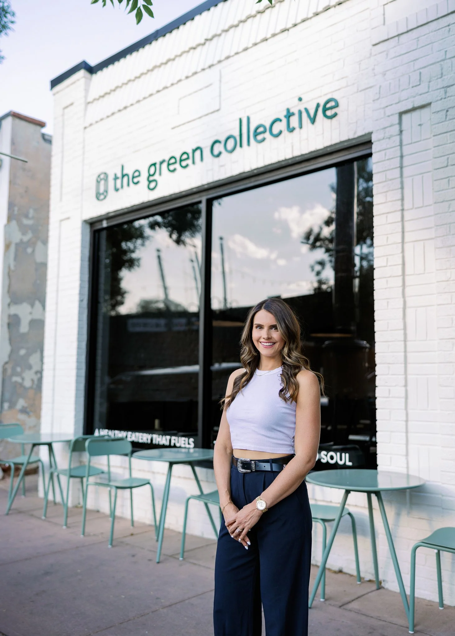 A smiling woman standing outside a storefront for 'the green collective', with teal chairs and tables outside on a sidewalk, storefront window reflecting a partly cloudy sky and trees.