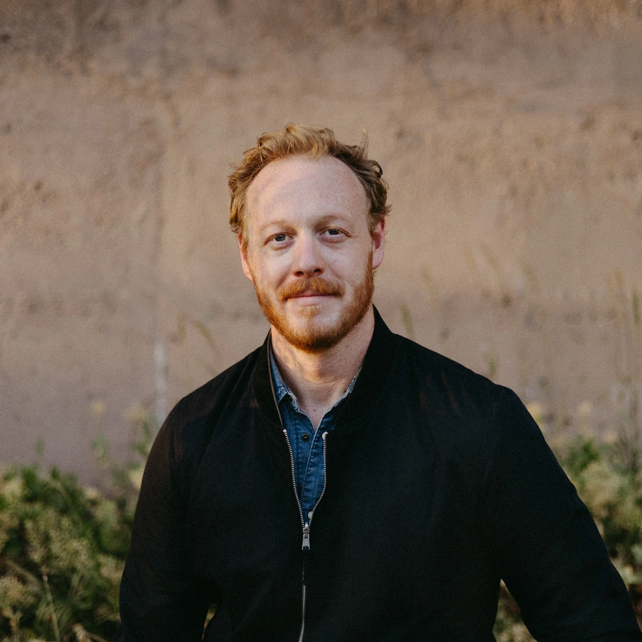 A man with red hair and beard wearing a black jacket and blue shirt outdoors.