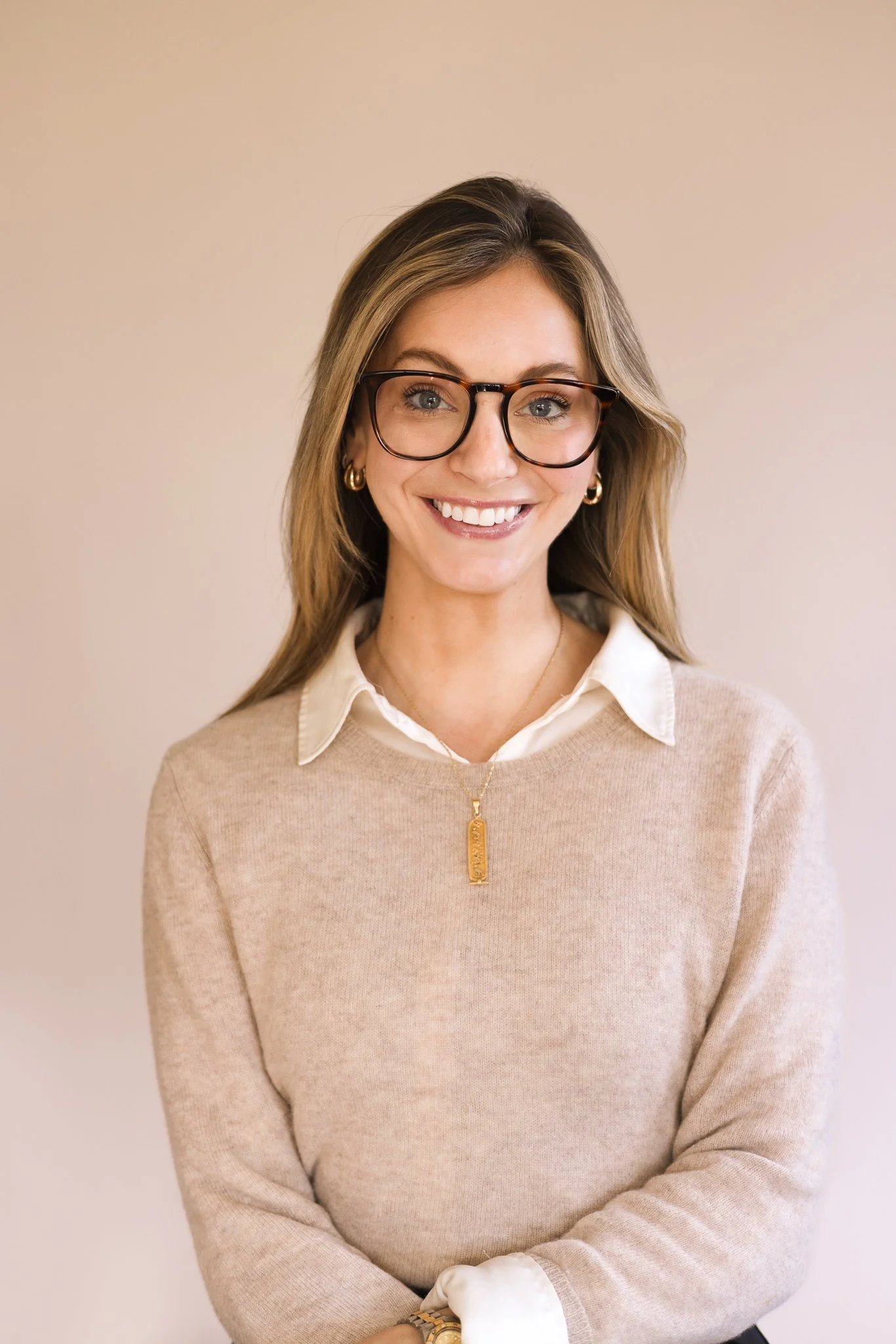 A woman with long blonde hair wearing glasses, gold hoop earrings, a beige sweater over a white collared shirt, a gold necklace, and a watch, smiling against a plain beige background.