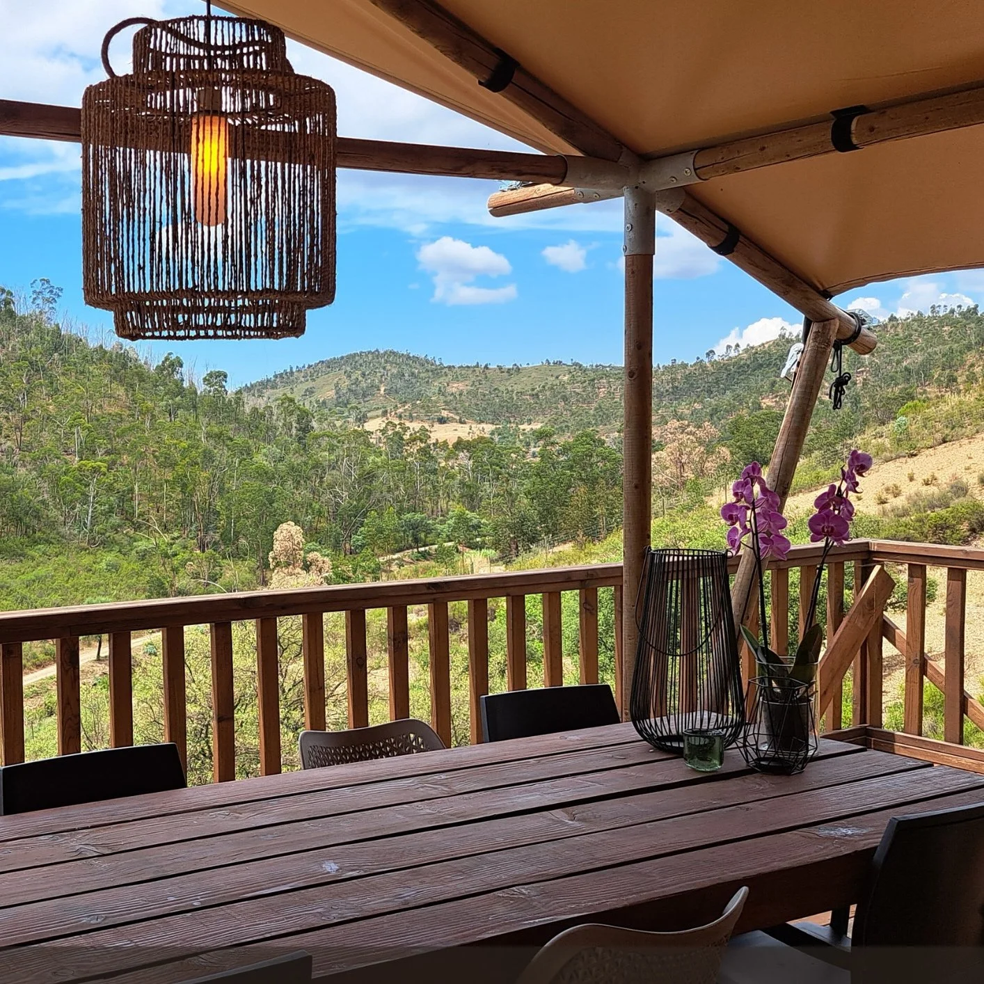 Wooden outdoor dining table on a balcony or porch with a view of green rolling hills, a wooden railing, purple orchids in a black wire basket, and a woven pendant light hanging from the ceiling.