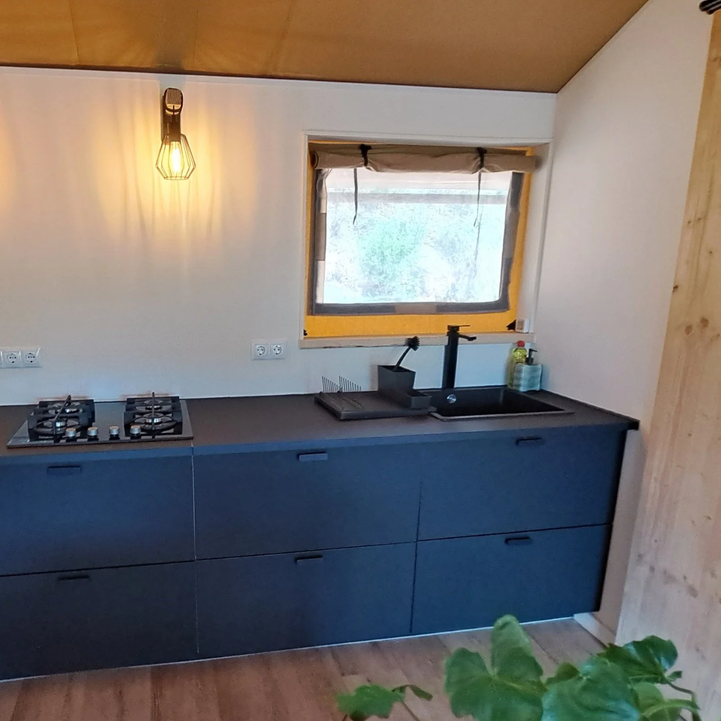 A modern kitchen with a black countertop, built-in stove, black sink, and a window above the sink. There is a warm industrial-style hanging light on the left and a wooden wall on the right. The window has a beige roller shade and the floor appears to