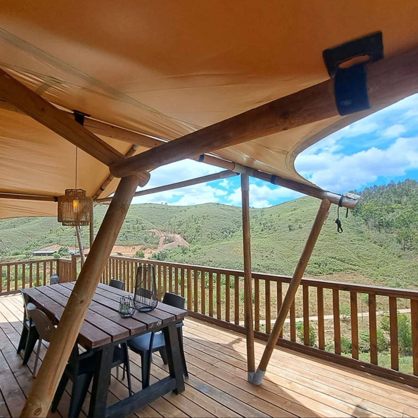 Outdoor wooden deck with a dining table, chairs, and a tent canopy overlooking green rolling hills and blue sky with clouds.