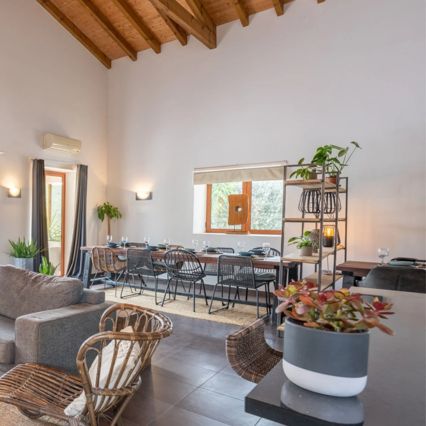 Living and dining room with high wooden ceiling, white walls, a large window, dining table with chairs, a sofa, and decorative plants.