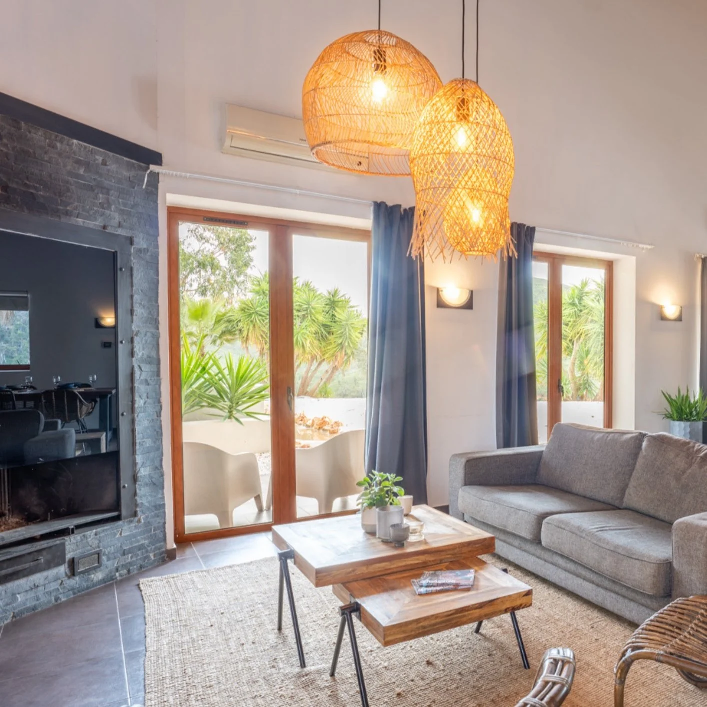 Cozy living room with a gray sofa, wooden coffee table, hanging rattan pendant lights, blue curtains, and sliding glass doors leading to an outdoor patio with green plants