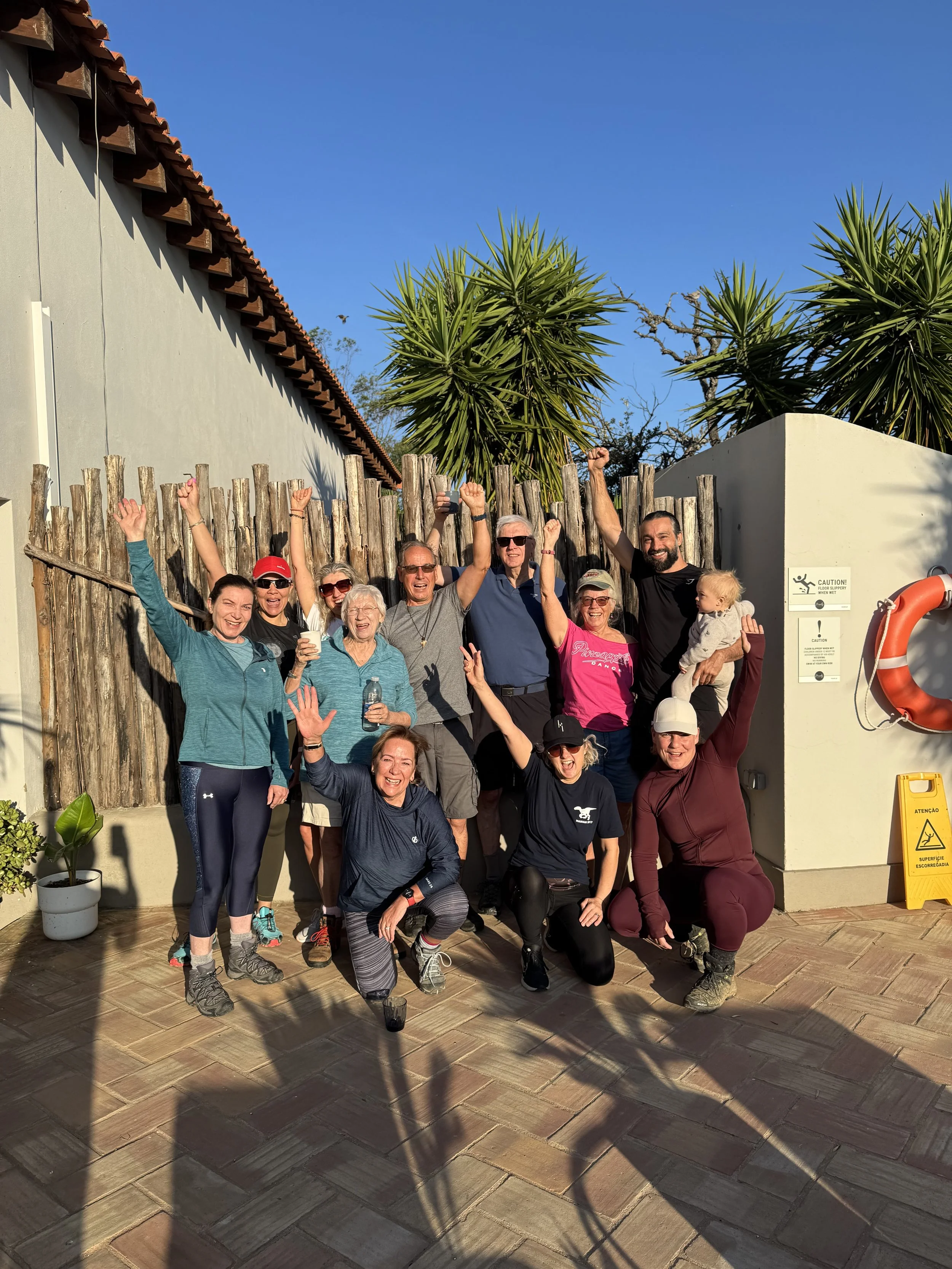Group of people outdoors celebrating, with some raising their arms, standing on a paved patio near a wooden fence, with palm trees and a blue sky in the background.