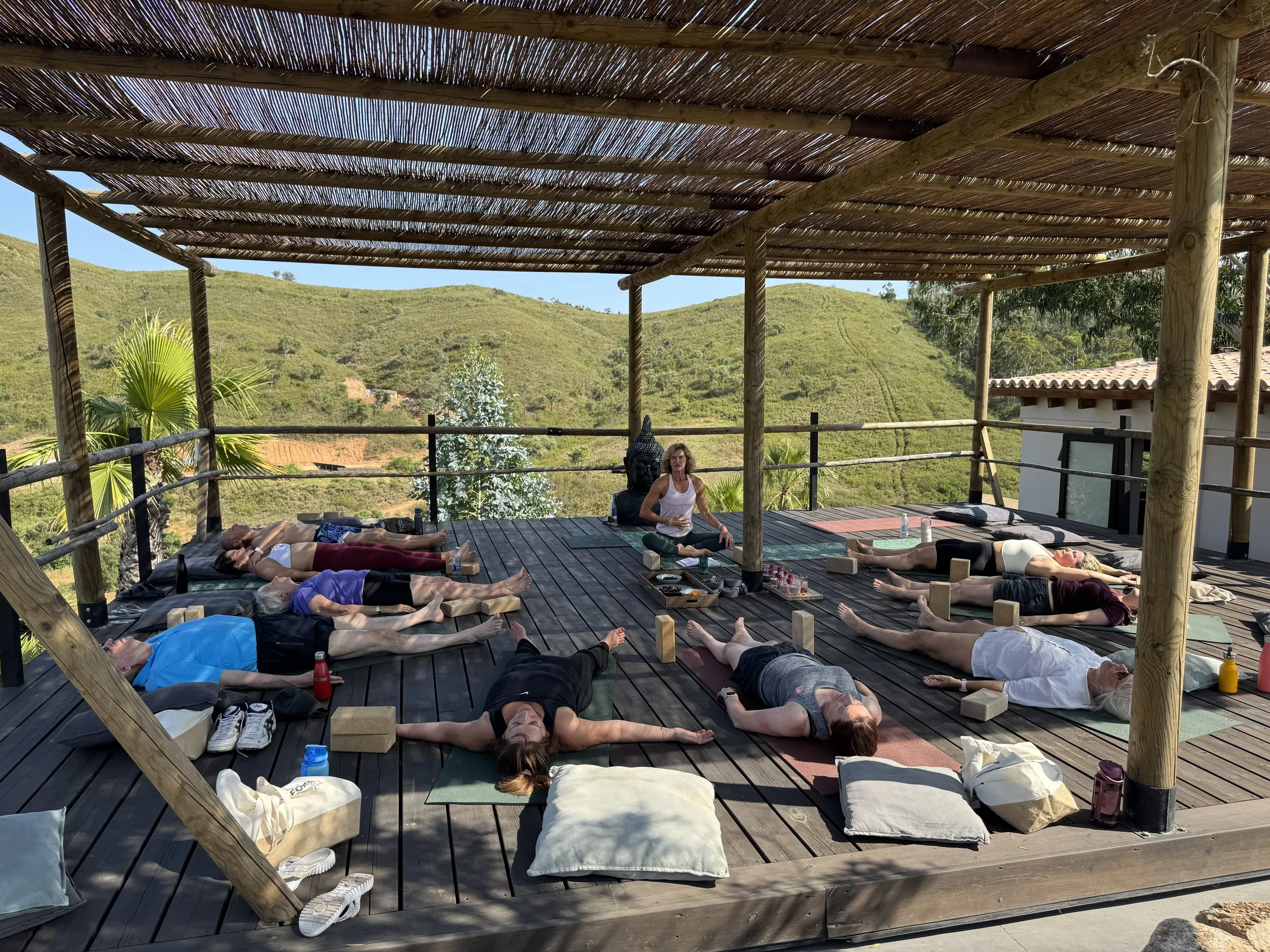 Group of people participating in an outdoor yoga or meditation class on a wooden deck with a thatched roof, surrounded by green hills and blue sky.