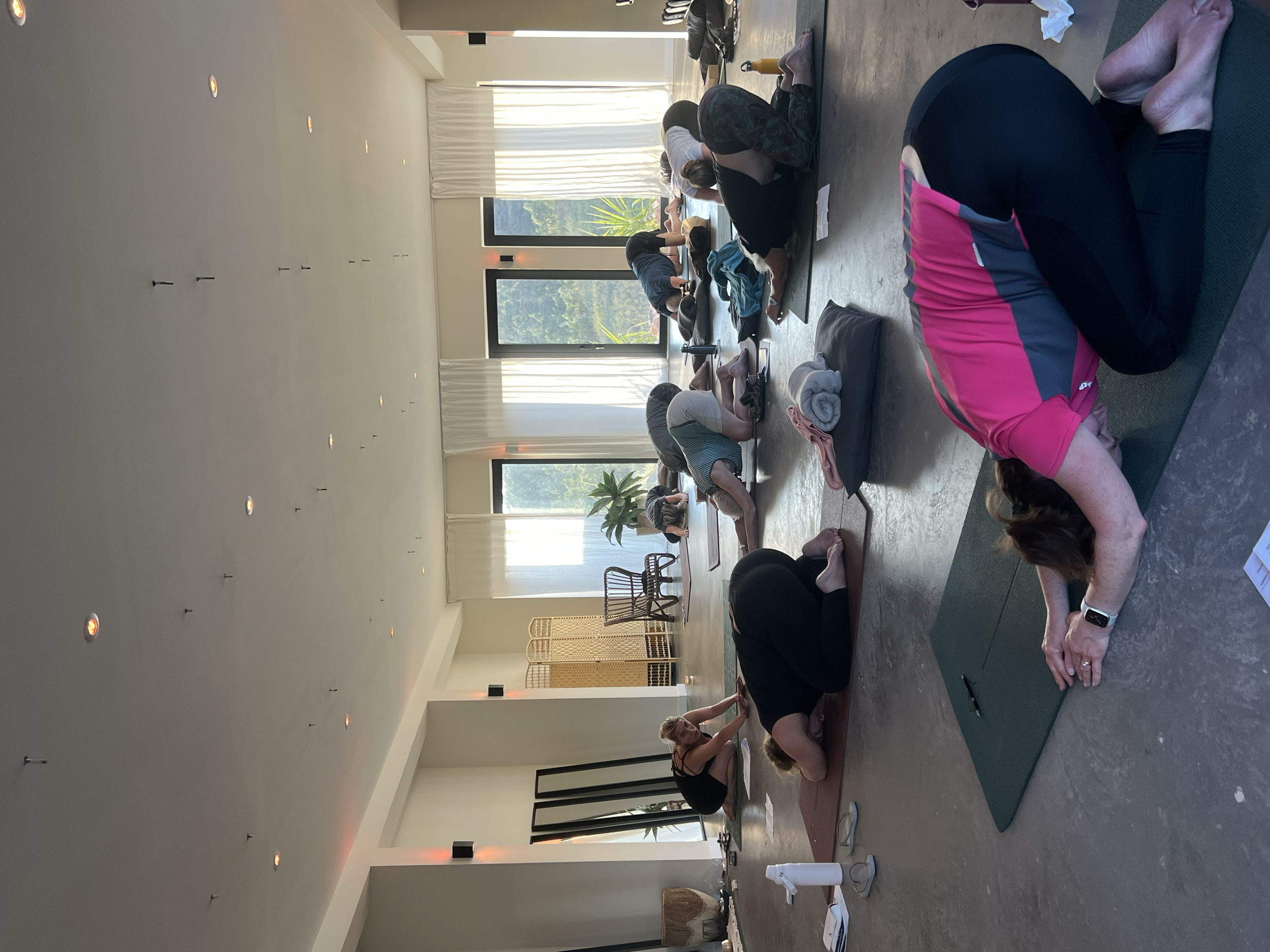 Group of people participating in a yoga class in a bright, spacious room with large windows, practicing various yoga poses on mats.