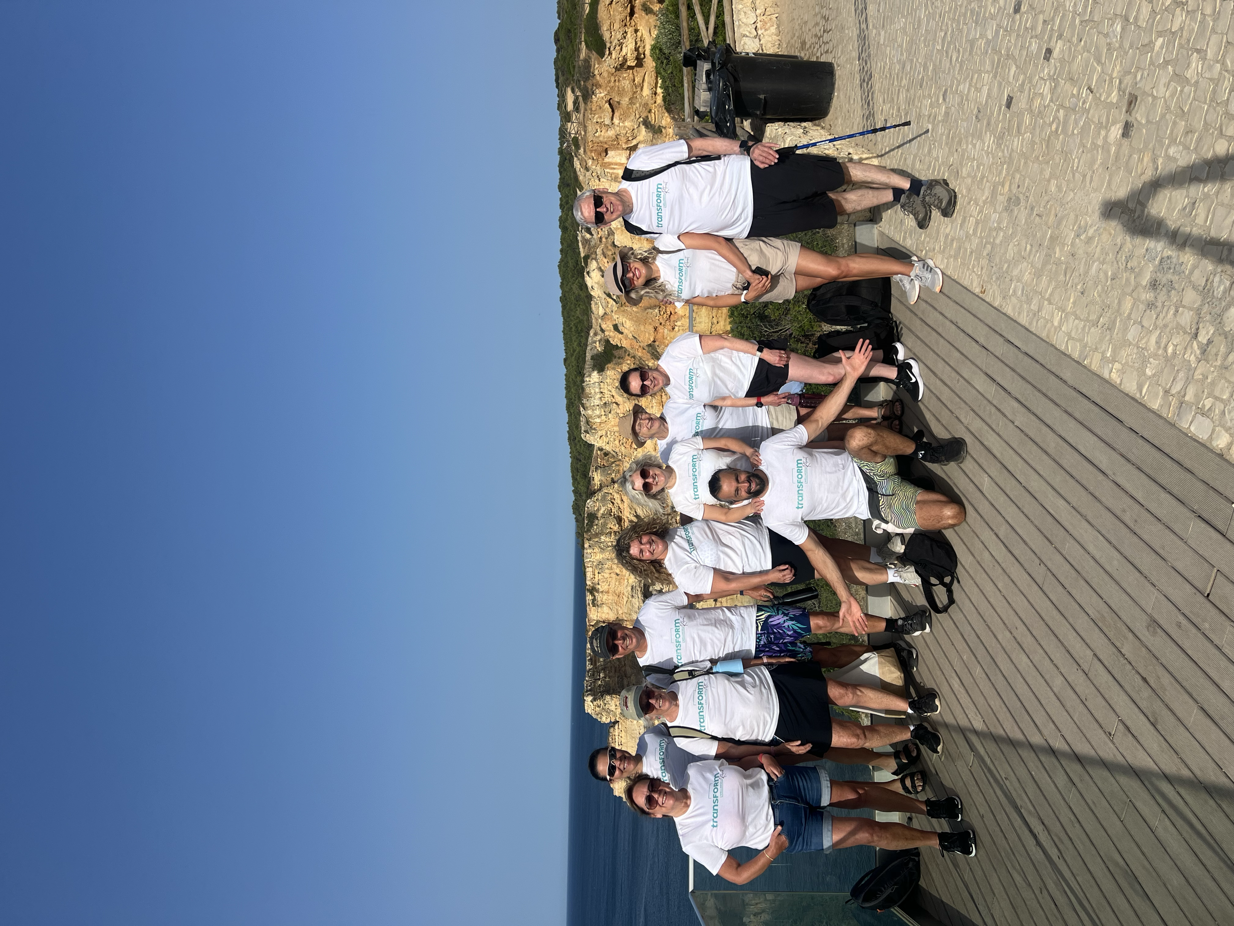 Group of eight people on a wooden dock beside the ocean, wearing white t-shirts, some with sunglasses, posing for a photo with a rocky landscape and blue sky in the background.