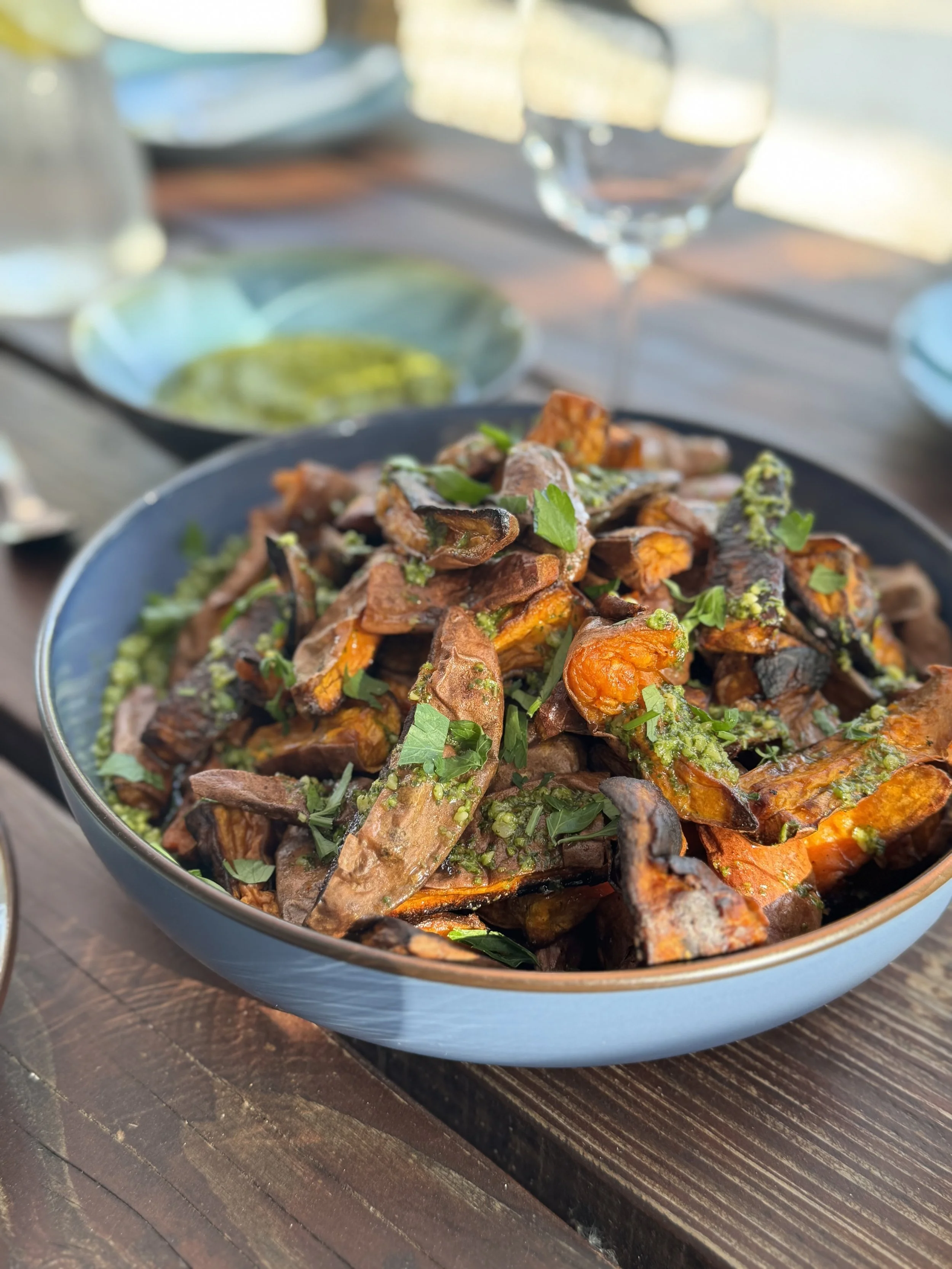 A bowl of roasted sweet potatoes and vegetables garnished with chopped herbs on a wooden table, with glasses of water and a small dish of green sauce in the background.
