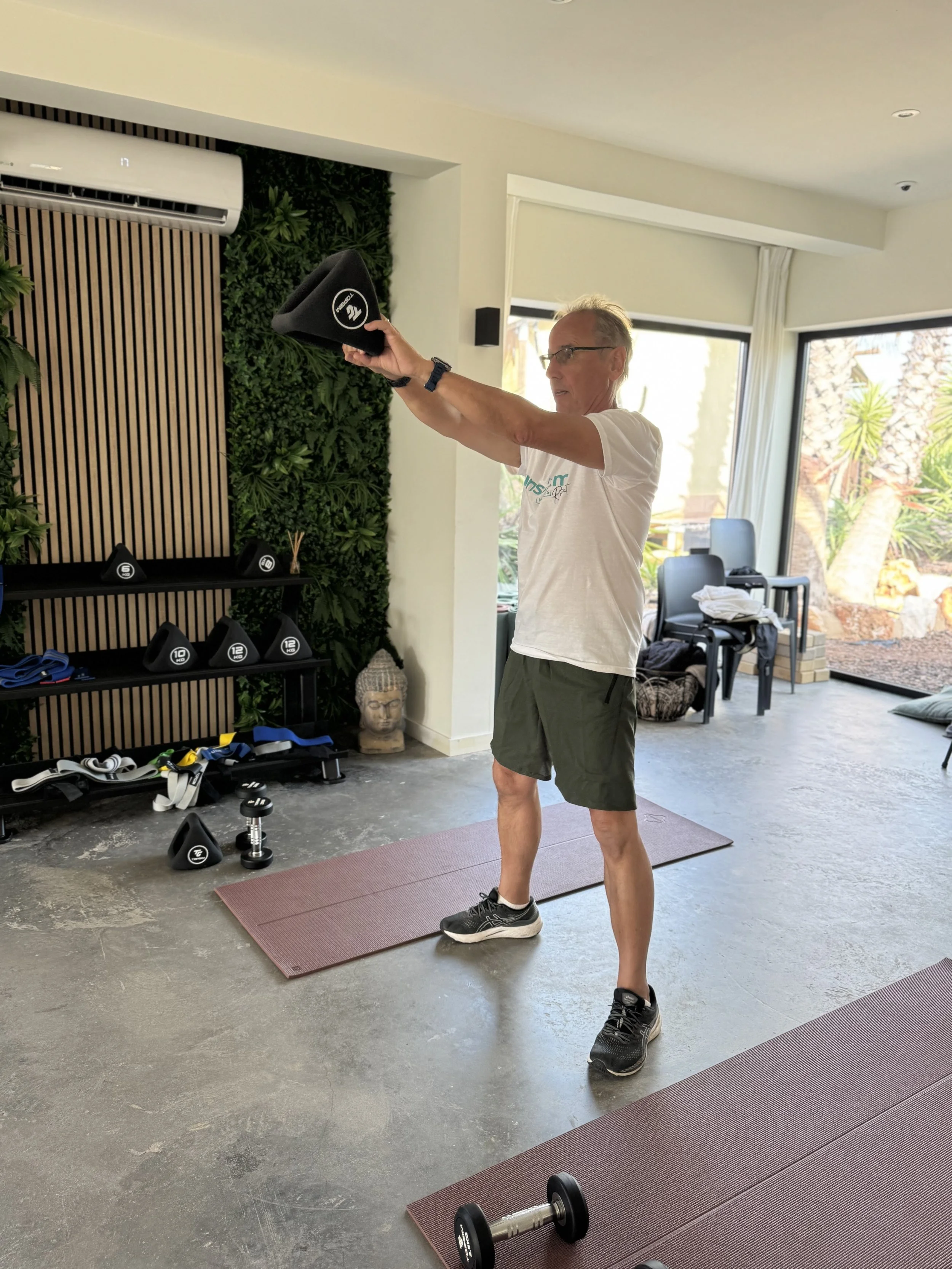 A man in a white t-shirt and black shorts is doing a workout with a weight horn in a home gym, standing on a yoga mat and lifting the horn with both hands. There are weights, resistance bands, and a shelf with dumbbells in the background, along with