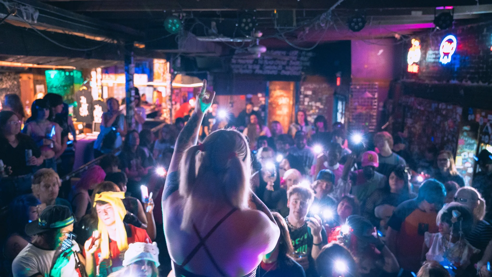 Crowd at a lively music venue with people taking photos and holding phones up, vibrating neon signs in the background, colorful stage lighting, exposed brick walls, and a woman with braids dancing in the foreground.