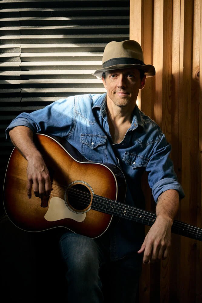Jason Mraz holding a guitar in front of a textured wall