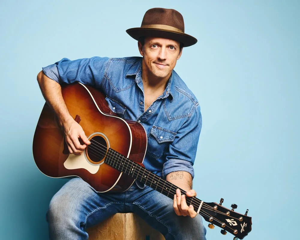 Jason Mraz holding a guitar in front of a blue background