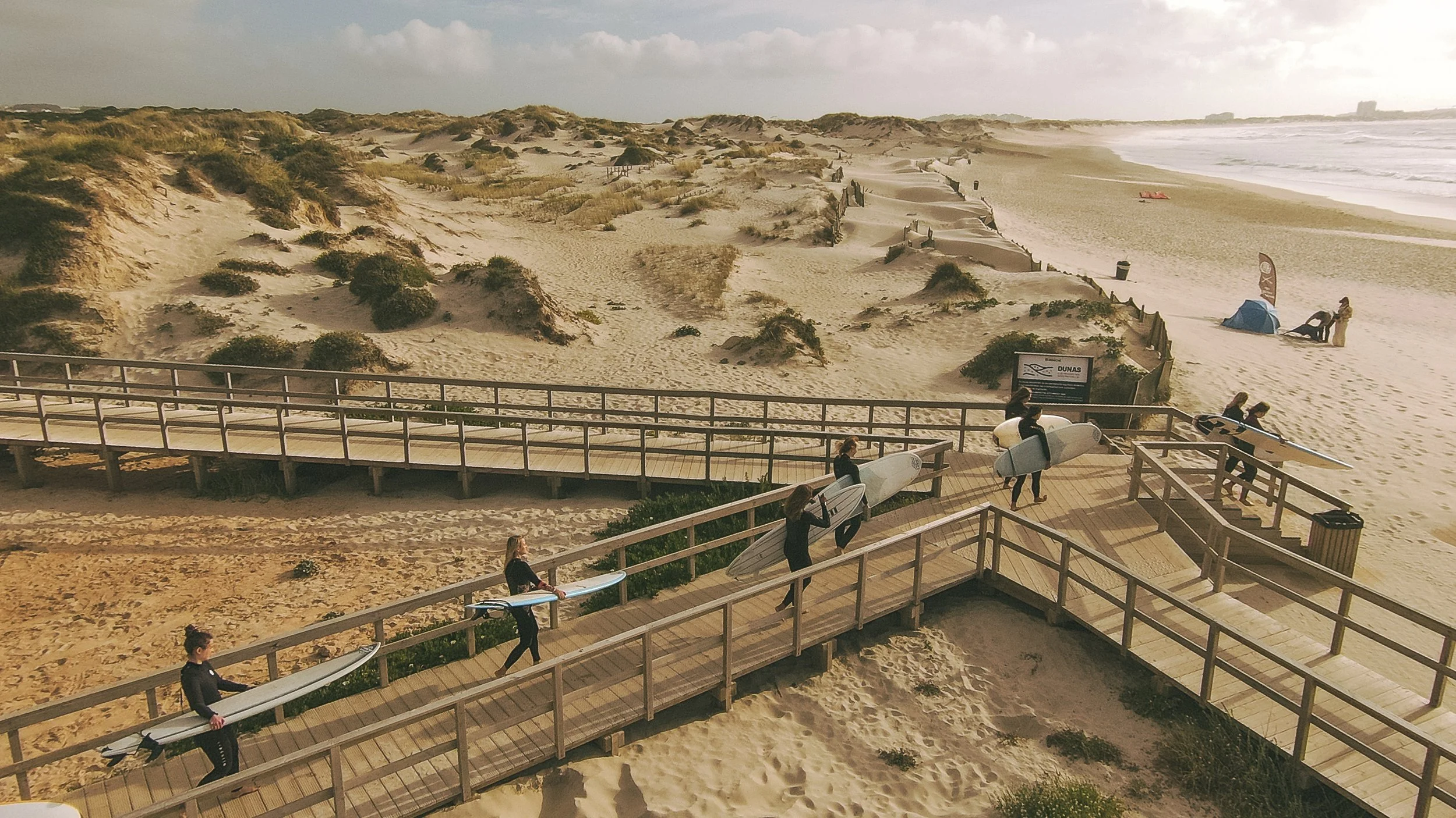 Beach scene showing a wooden boardwalk with surfers carrying surfboards, sandy dunes with sparse vegetation, and the ocean with waves in the background.