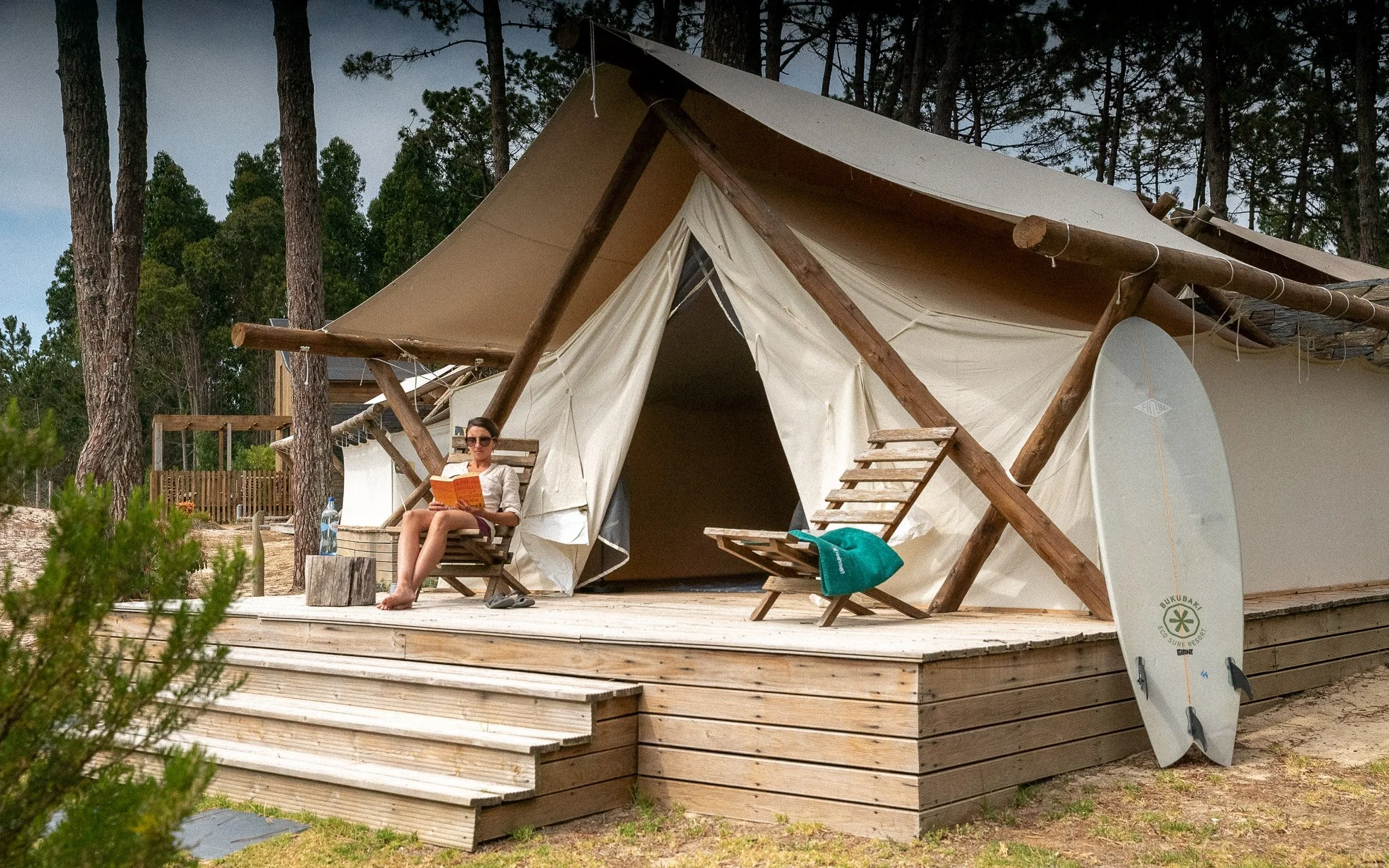 A woman sitting on a wooden deck porch outside a large canvas and wooden structure tent, reading a book with a bottle of water beside her, surrounded by tall trees.