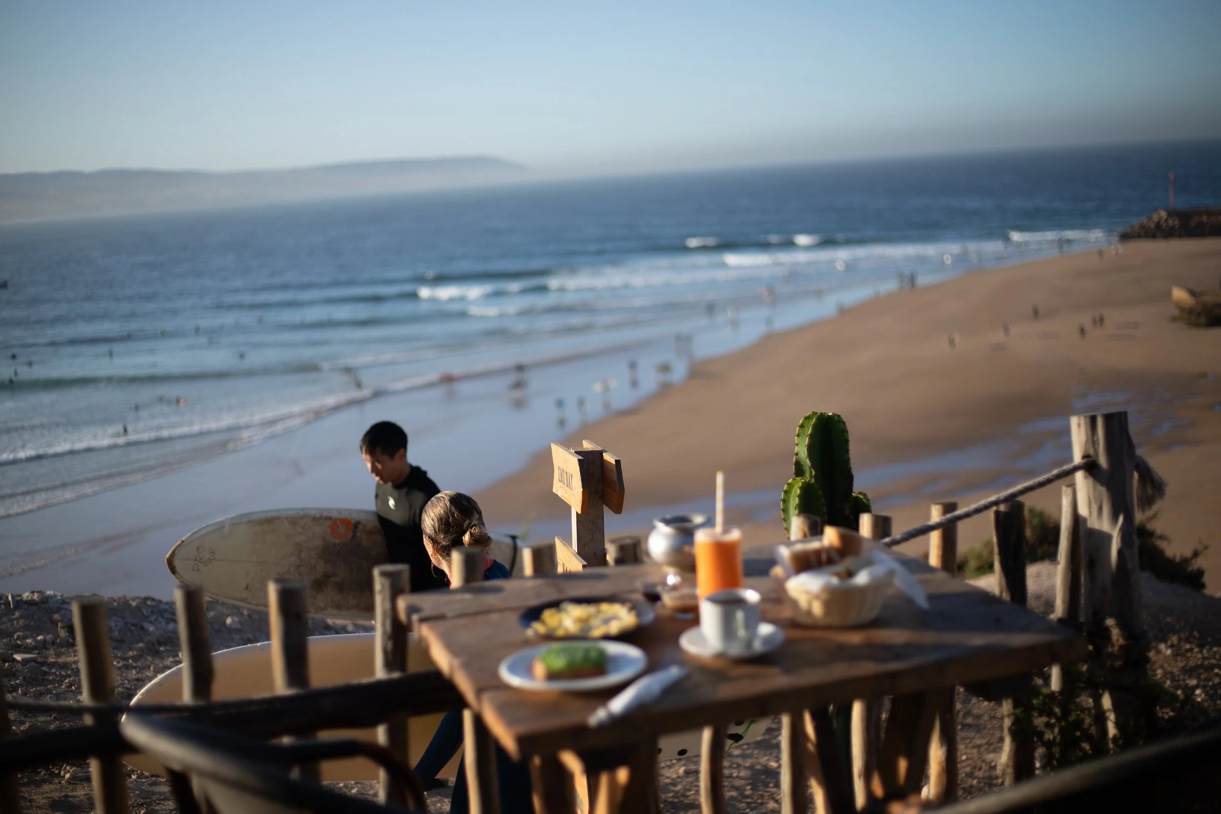 A beach scene with a table set with breakfast foods in the foreground, two children with surfboards near the dunes, and the ocean with people swimming and surfing in the background.