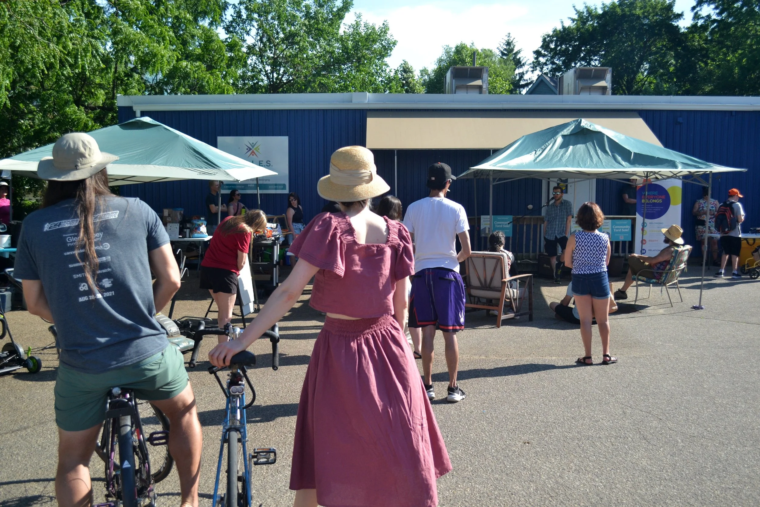 Outdoor community event with people waiting in line, seated, and standing in front of a blue building, with two umbrellas providing shade, and trees in the background.