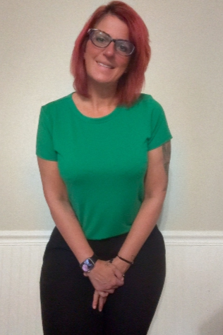 Woman with red hair, glasses, wearing a green t-shirt and black pants, smiling, standing in front of a light-colored wall.