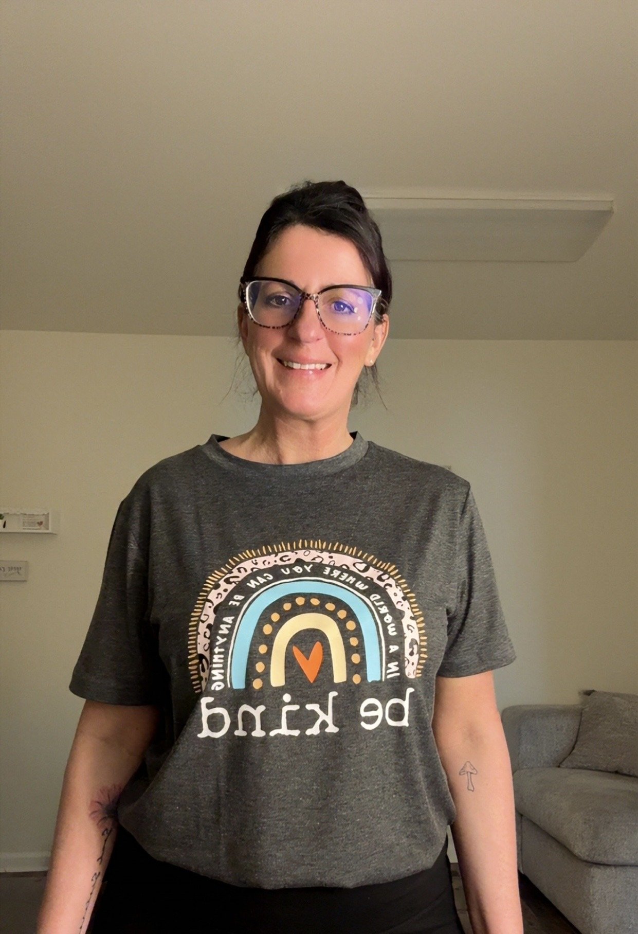 A smiling woman with short red hair and glasses, wearing a leopard print vest over a white shirt, standing against a plain light-colored wall.