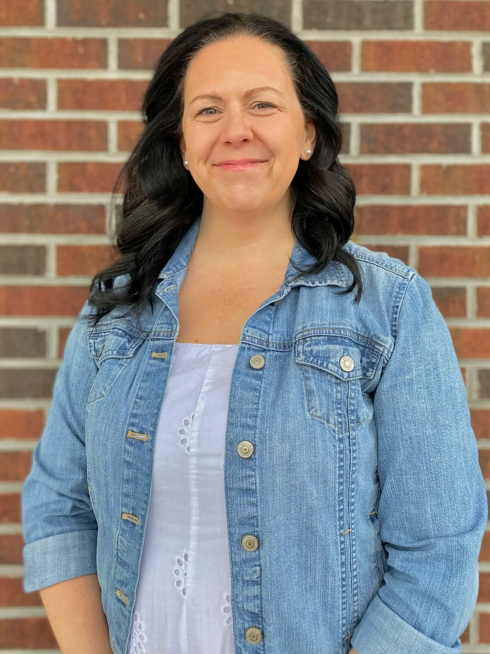 Woman wearing a jean jacket and white dress standing in front of a shingle wall