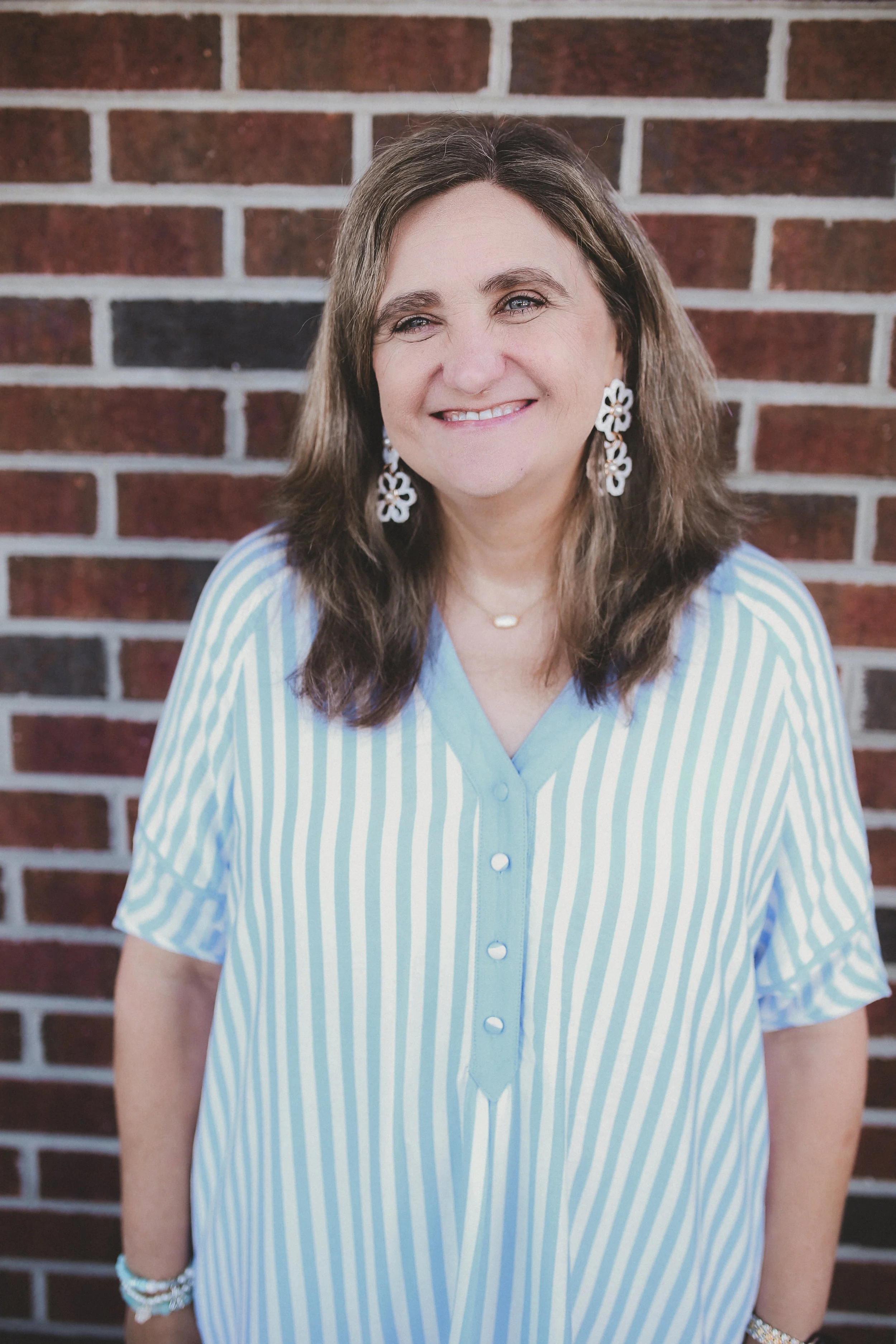 A woman smiling in front of a brick wall, wearing a blue dress with white floral patterns, with shoulder-length wavy brown hair.