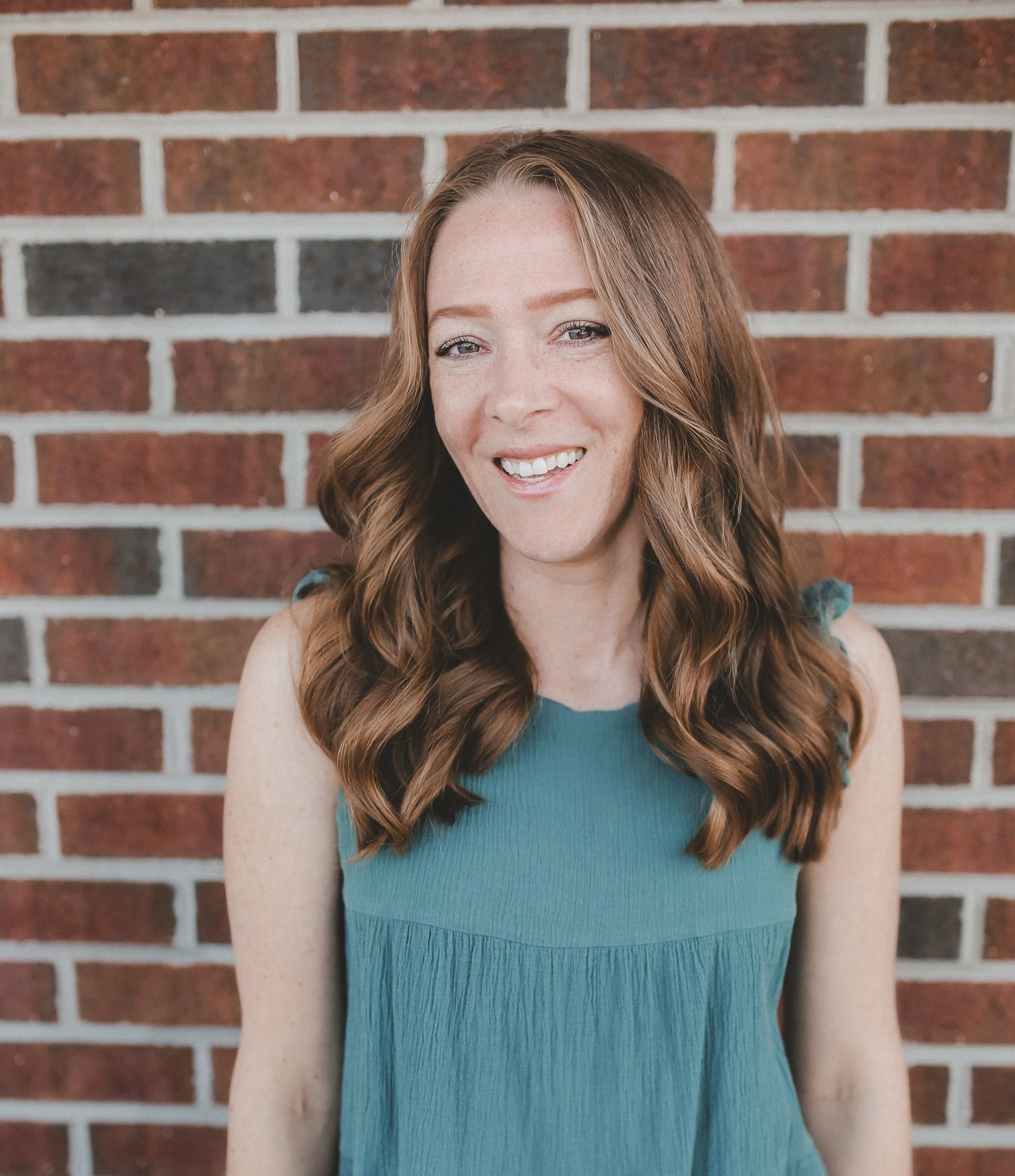 Woman with long red hair wearing a white sleeveless top, turquoise pants, layered necklace, and a smartwatch, standing in front of a light-colored shiplap wall.