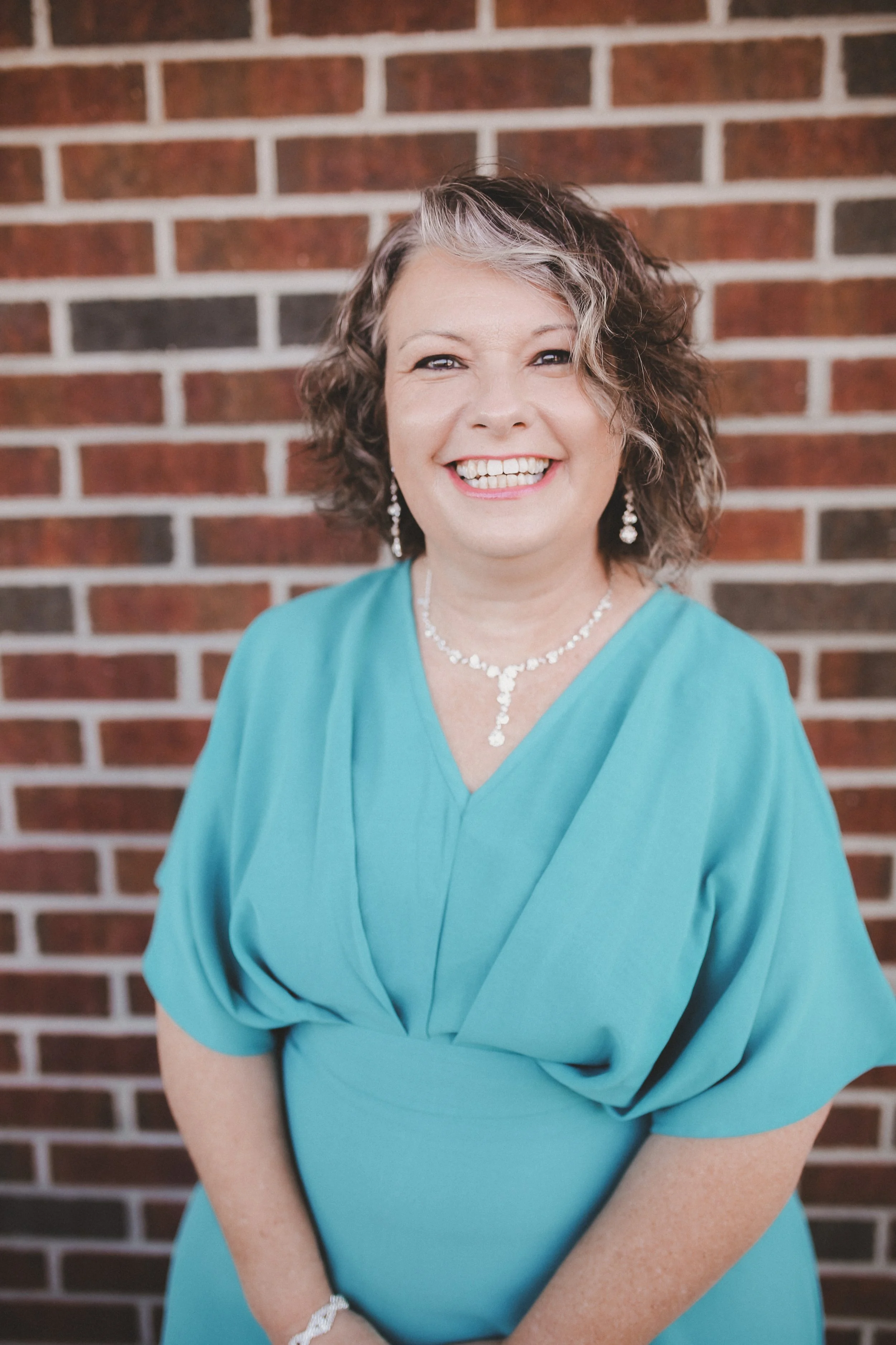 Woman wearing a green dress and gold hoop earrings, smiling in front of a white shiplap wall.
