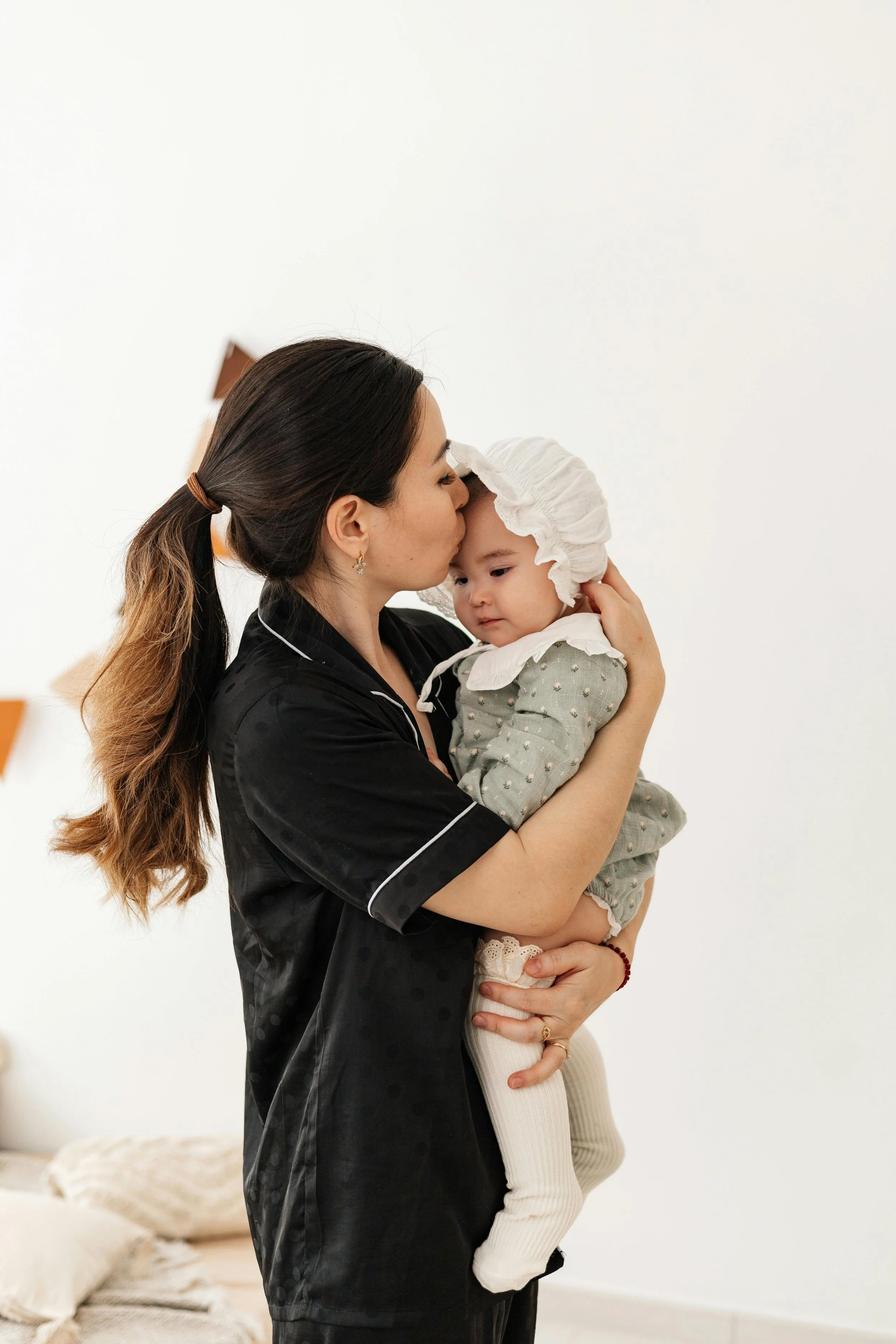 Woman in black clothing kissing a baby on the forehead, the baby wearing a bonnet and green outfit with white tights, in a light-colored room.
