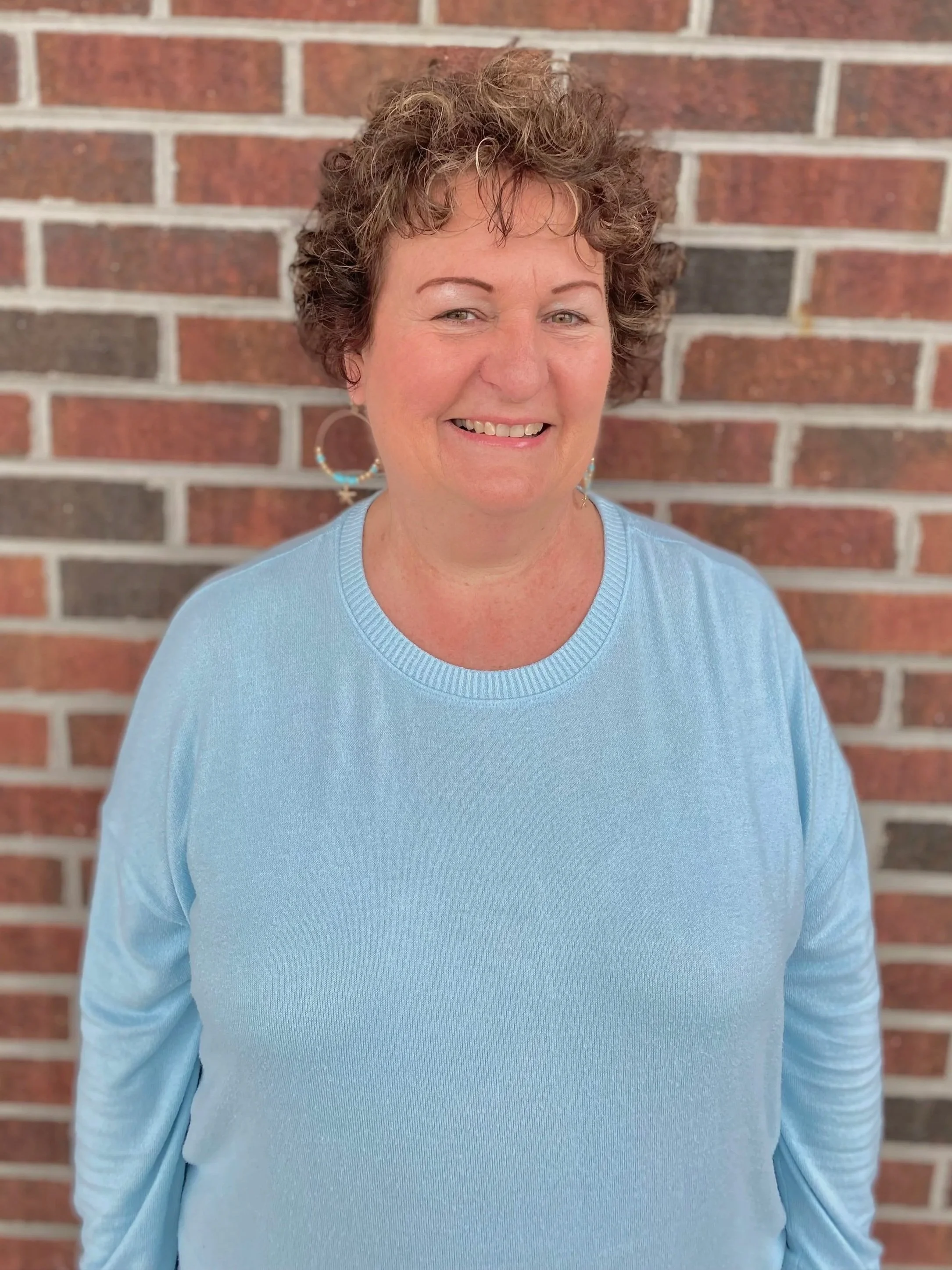 Person wearing a light blue blouse with a starfish necklace, standing against a wood-paneled wall.