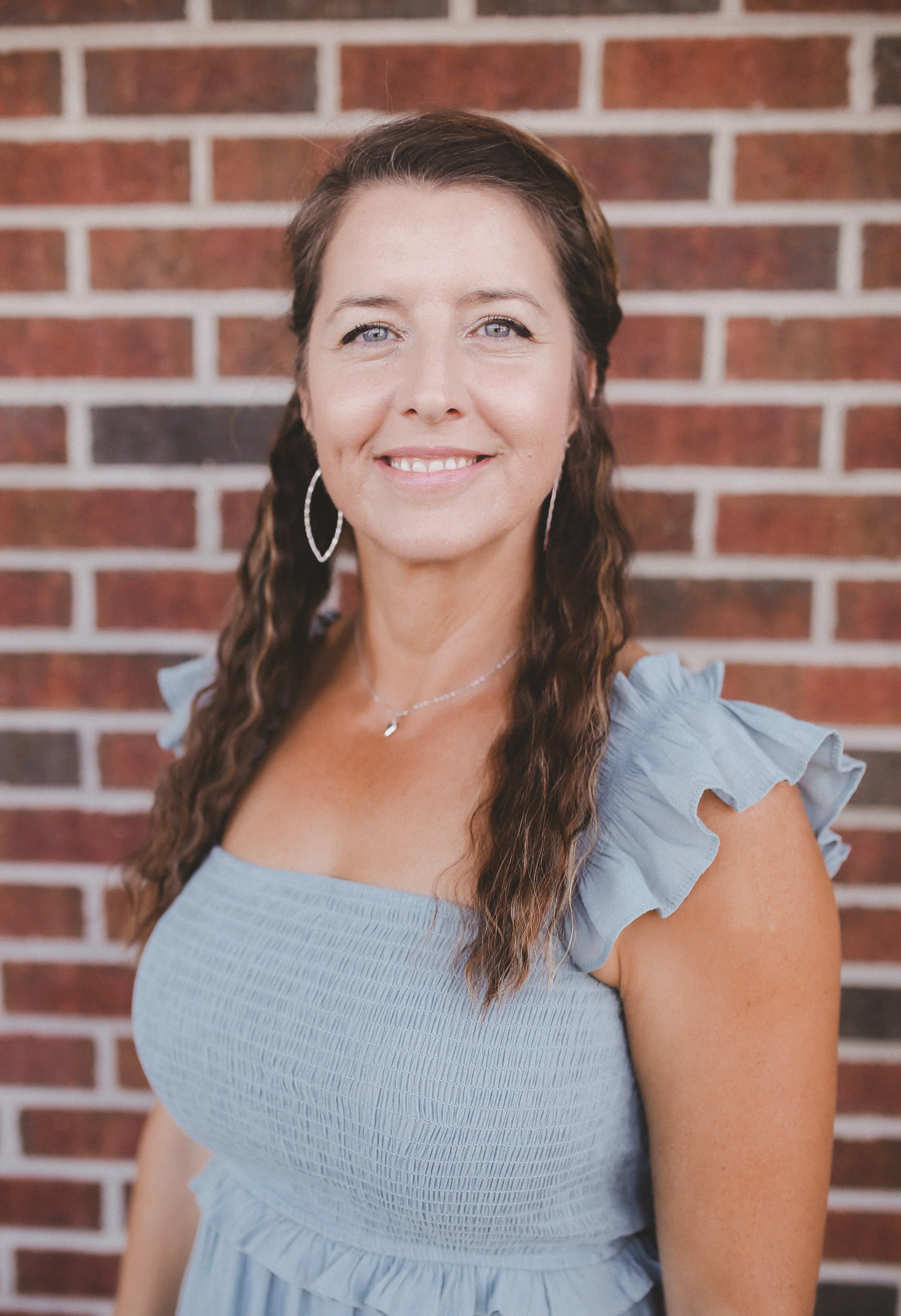 Portrait of a smiling woman with brown hair, wearing a white jacket, a turquoise top, a turquoise pendant necklace, and earrings, standing in front of a brick wall.