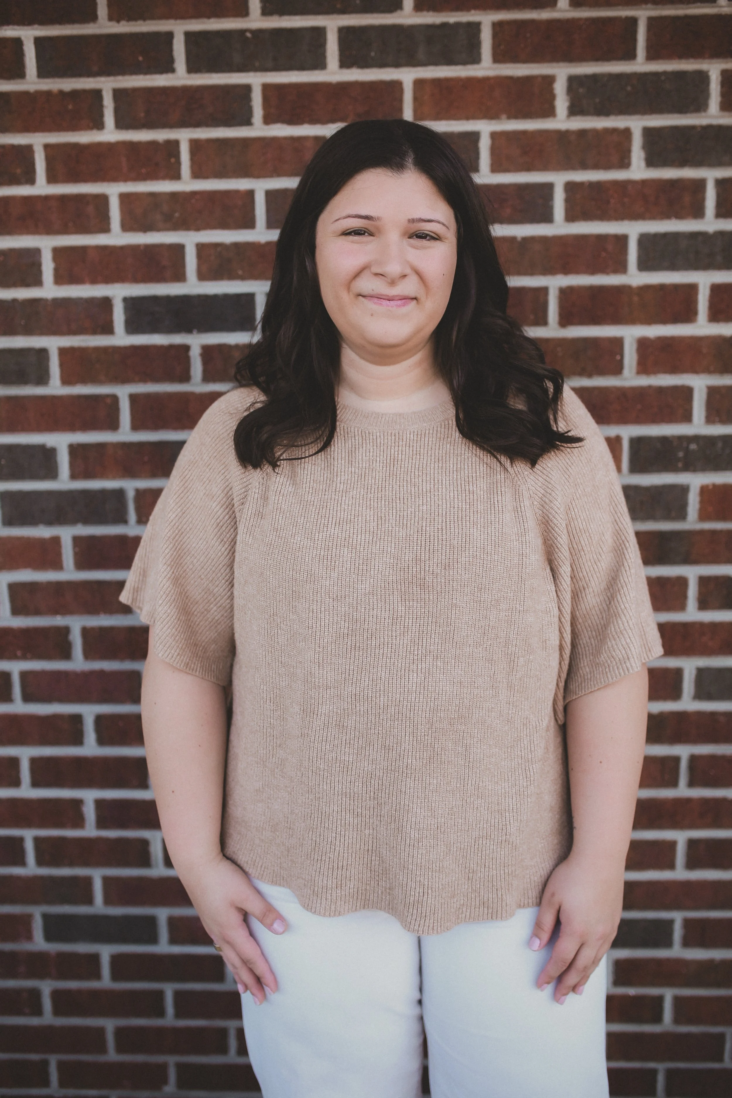 Woman in green top and white pants standing against a shingle wall, smiling.